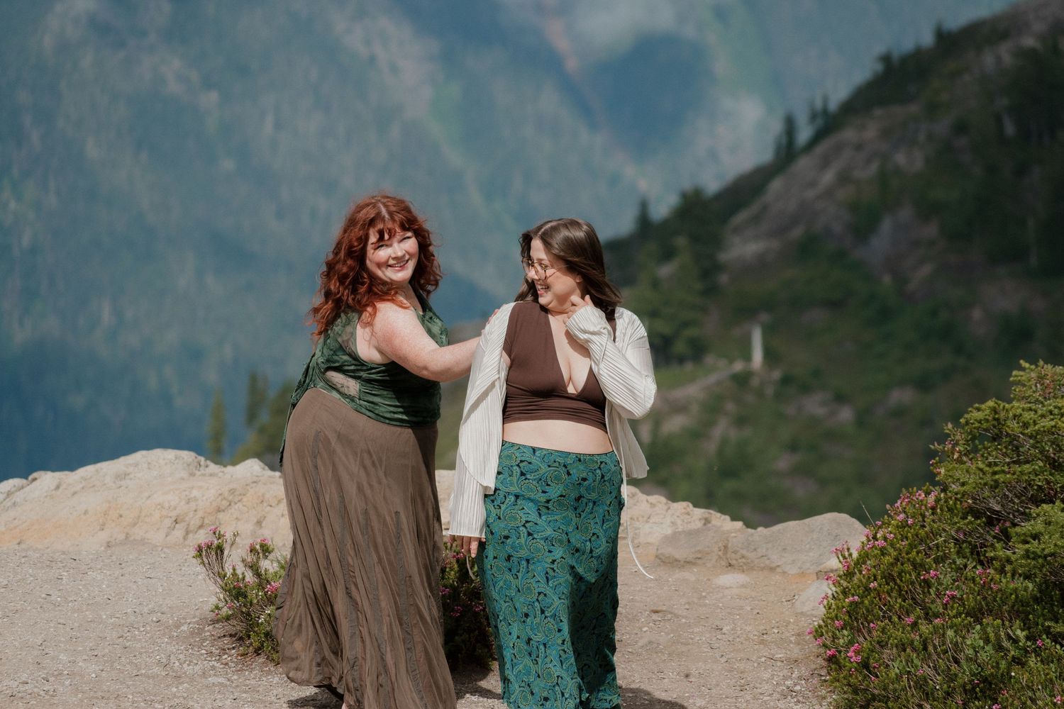 Two women laughing and walking together on rocky mountain overlook with scenic valley views.