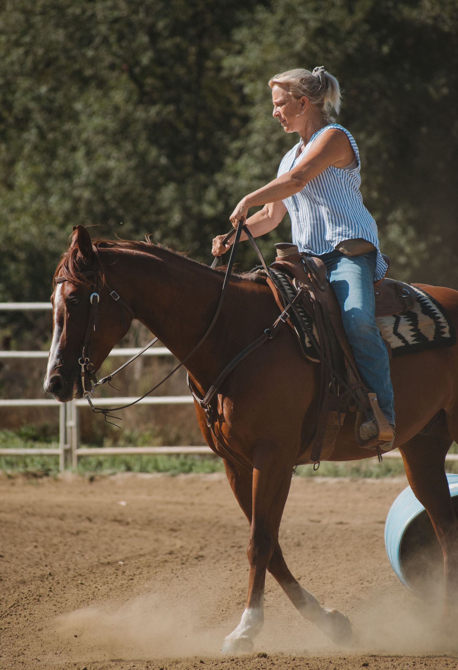 Blonde woman riding a chestnut horse in a sandy arena, wearing jeans and a blue top on a sunny day.