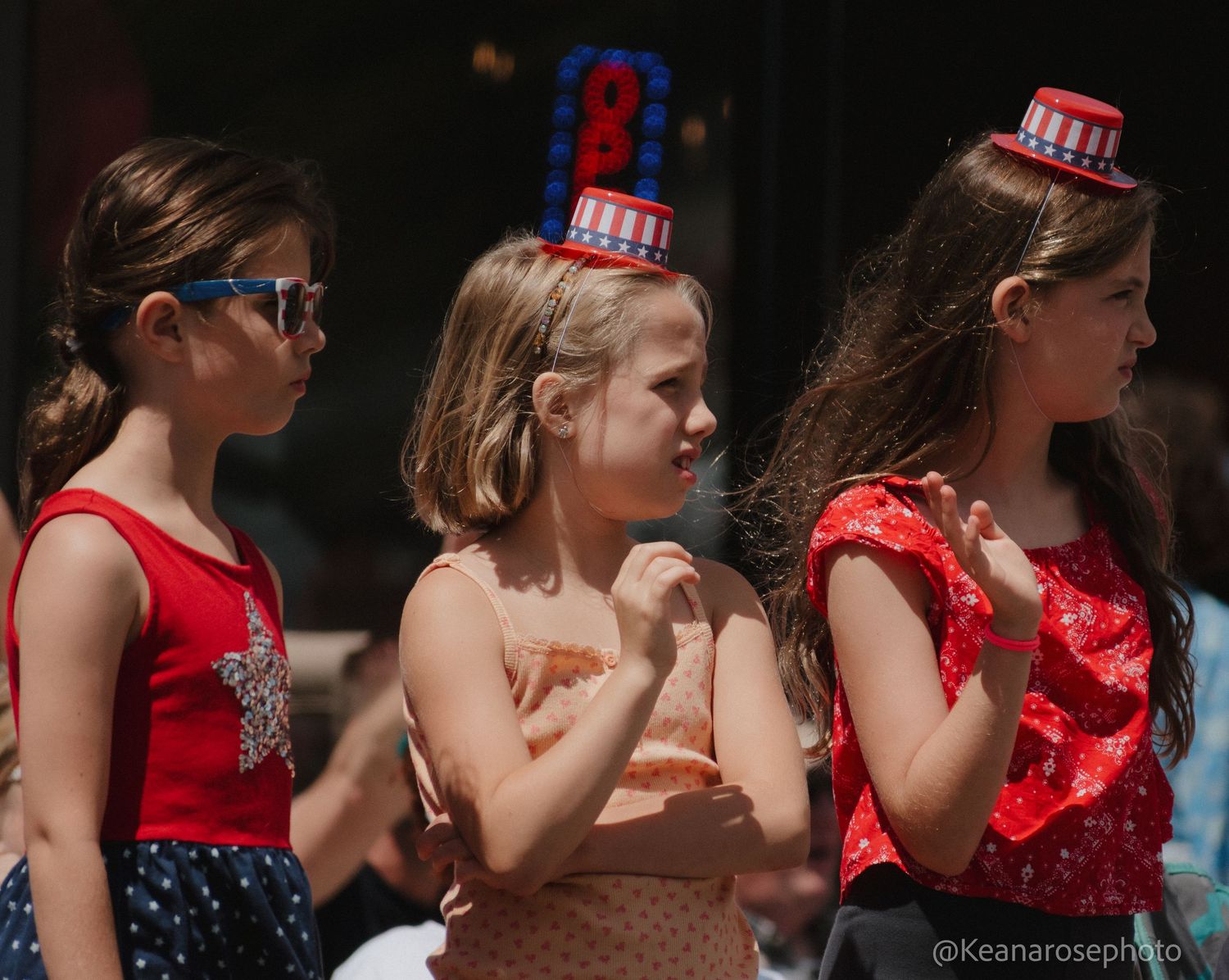 Three young girls in patriotic outfits wearing festive hats enjoy a sunny outdoor Independence Day parade.