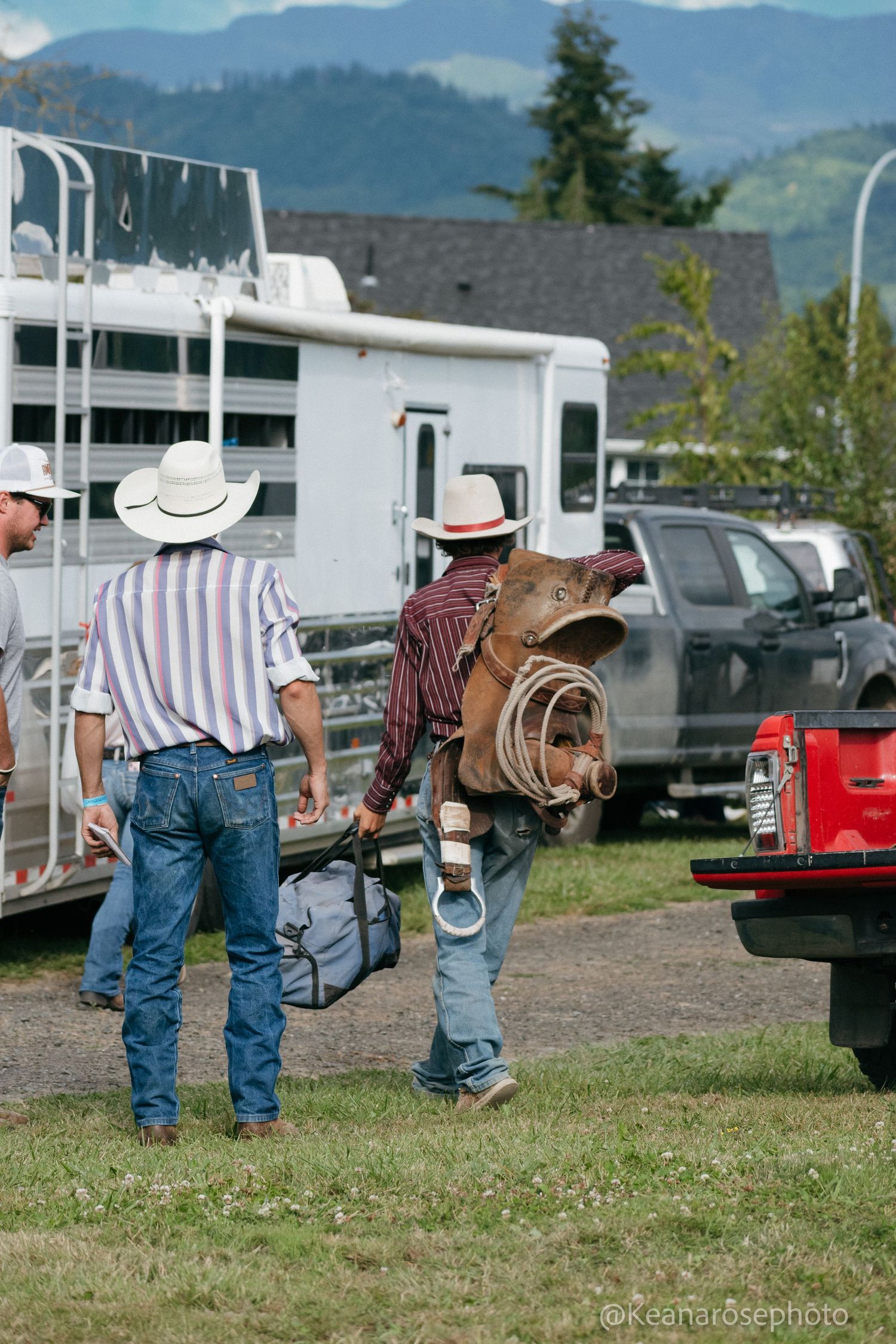 Two cowboys in white hats carry saddle and gear across grassy field near horse trailers and trucks.