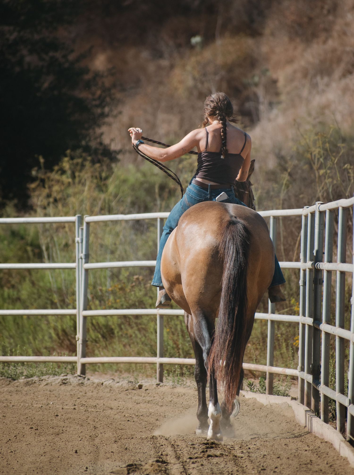 Woman riding brown horse in outdoor arena with braided hair and jeans.