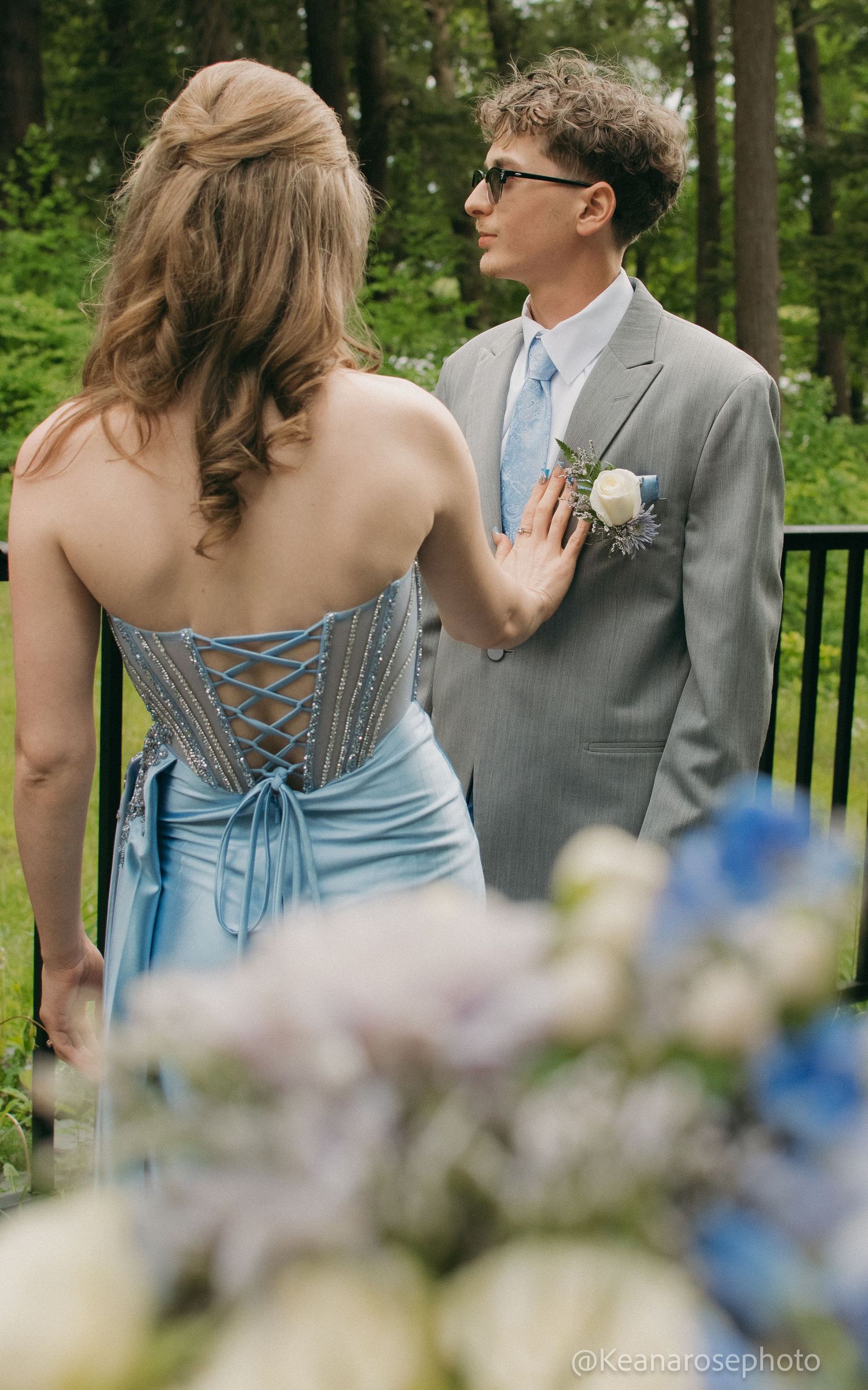 A couple in prom attire, blue gown and gray suit with boutonniere, pose together outdoors near greenery.