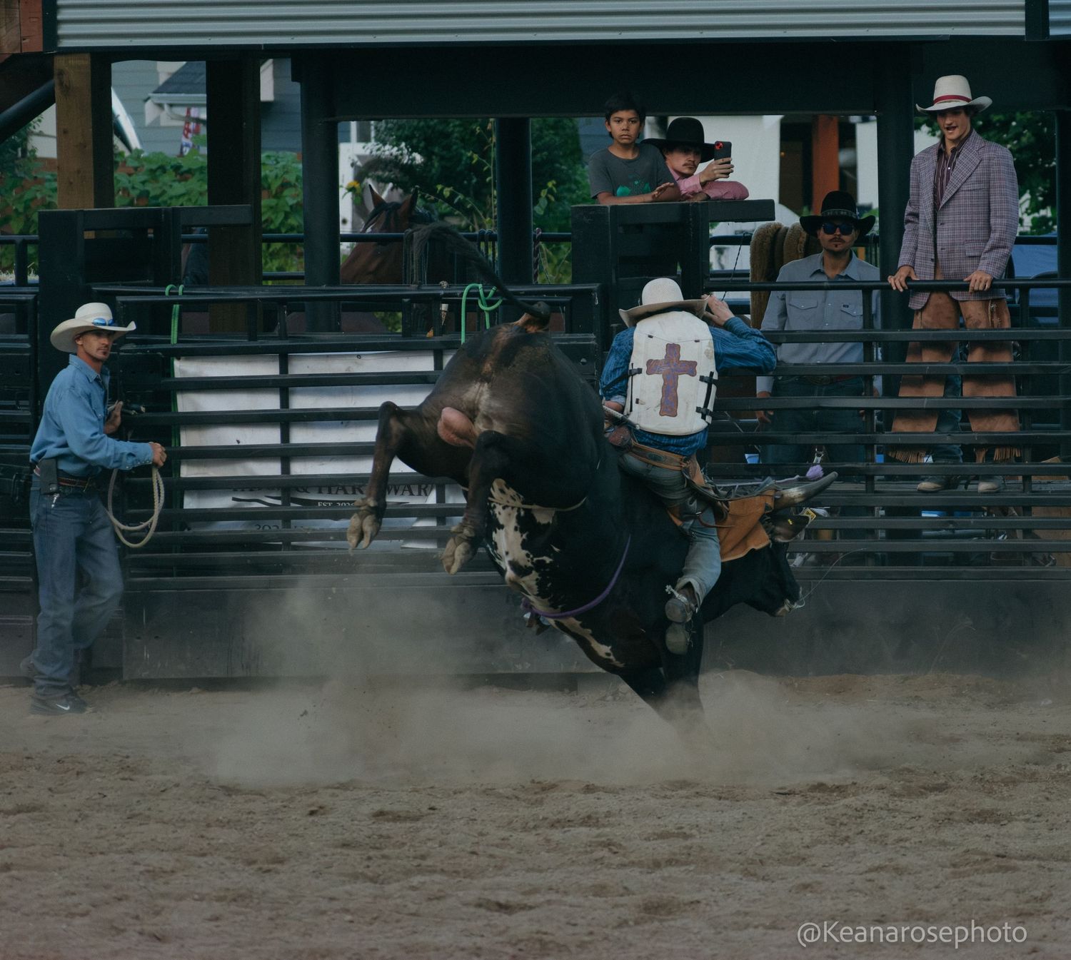 A cowboy rides a bucking black bull while a rodeo clown stands nearby in the arena.