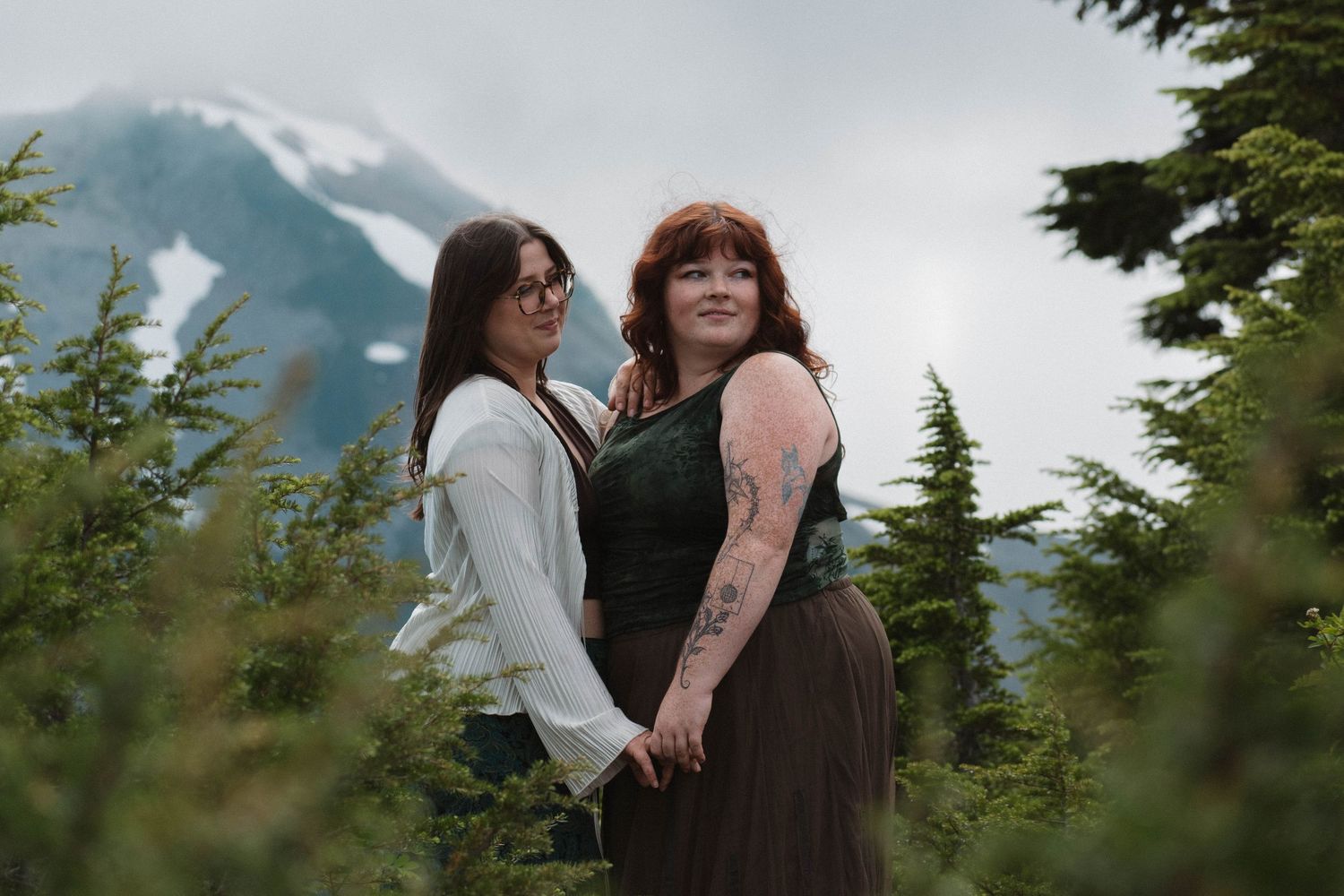 Two women hold hands and pose together among evergreen trees with a snow-capped mountain backdrop.