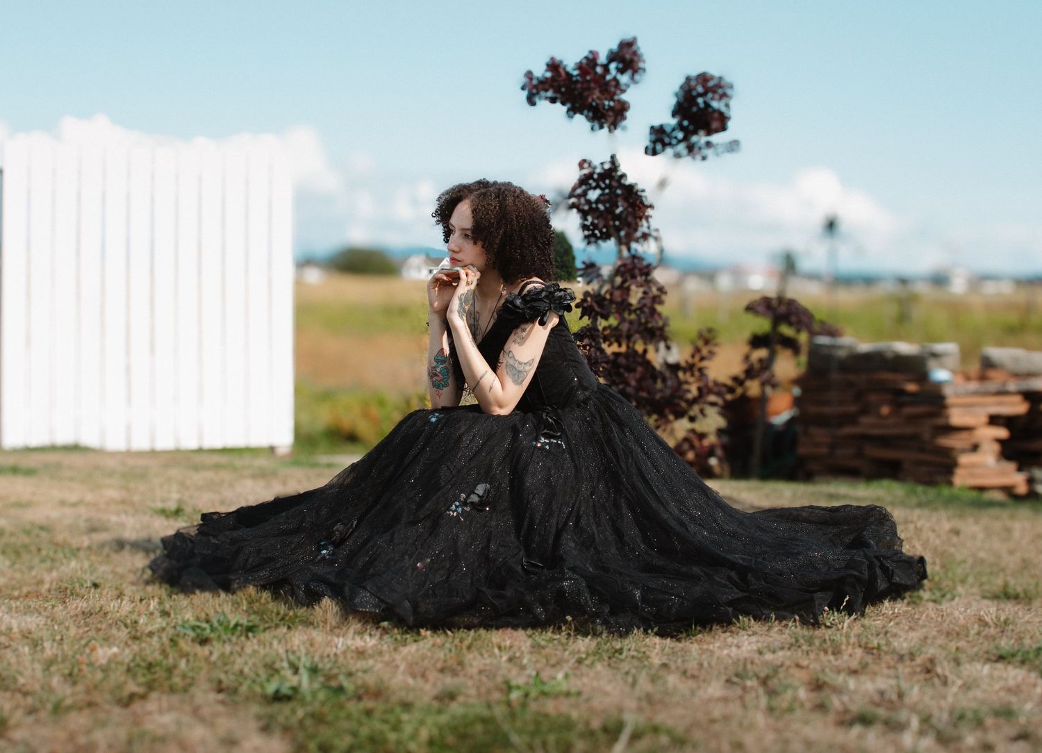 Woman in elegant black ball gown sitting gracefully on grass in outdoor rustic setting.