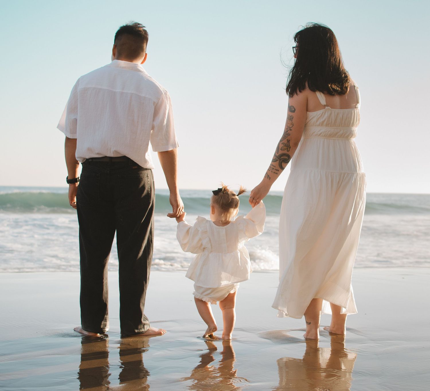 Family of three holding hands on a sandy beach, parents and toddler in white outfits facing ocean waves.