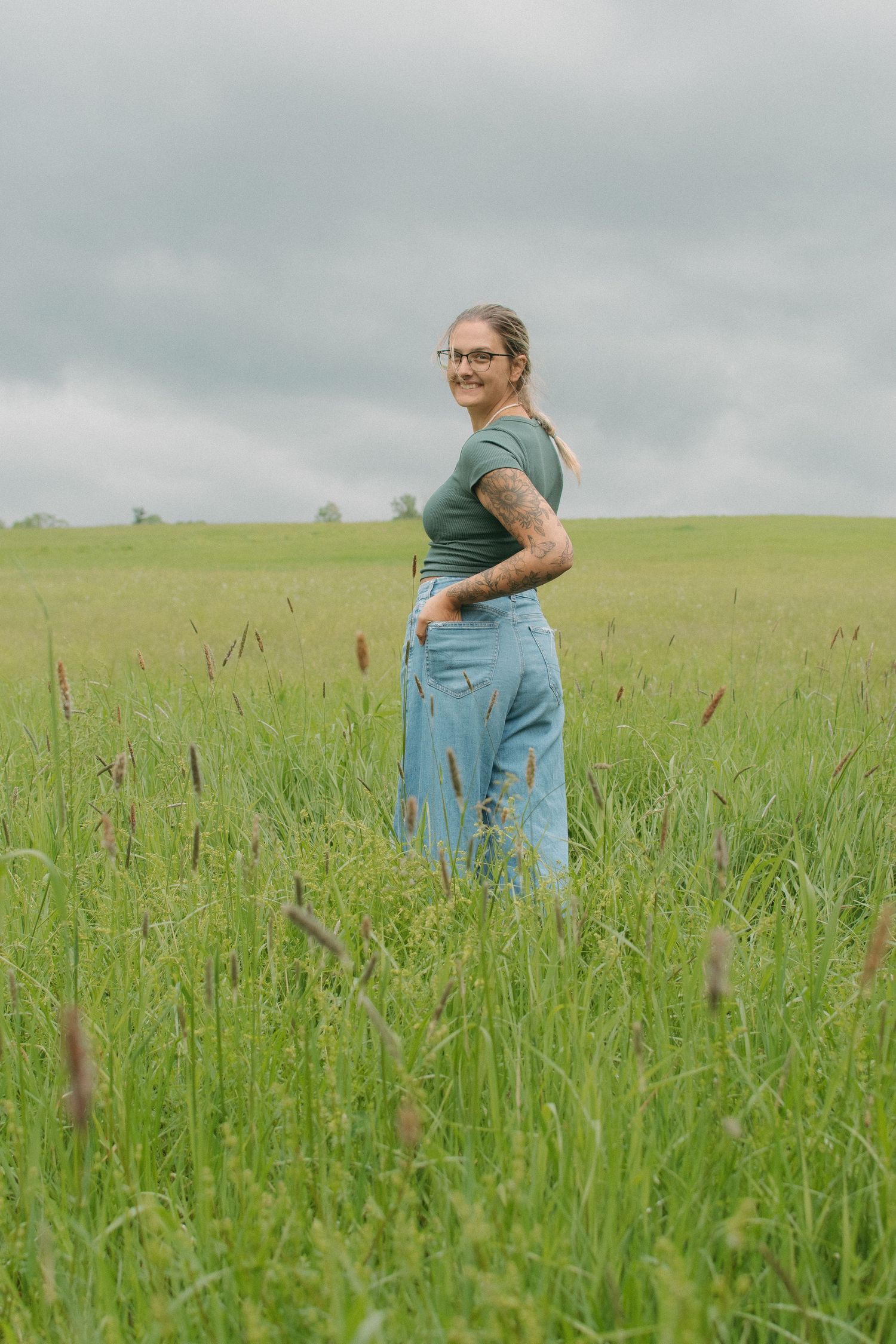 Woman with tattoos in green top and jeans standing in a lush green field under cloudy skies.
