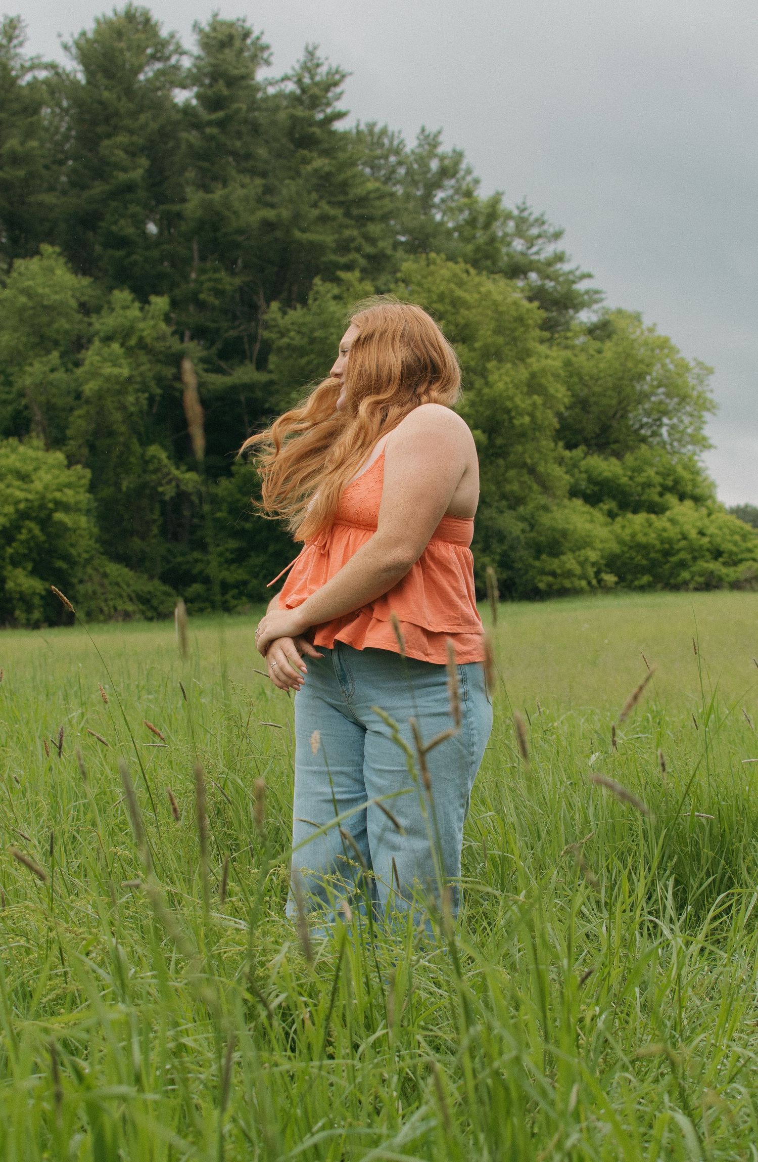 A woman with long blonde hair in an orange top and jeans stands in a lush green meadow with trees in the background.
