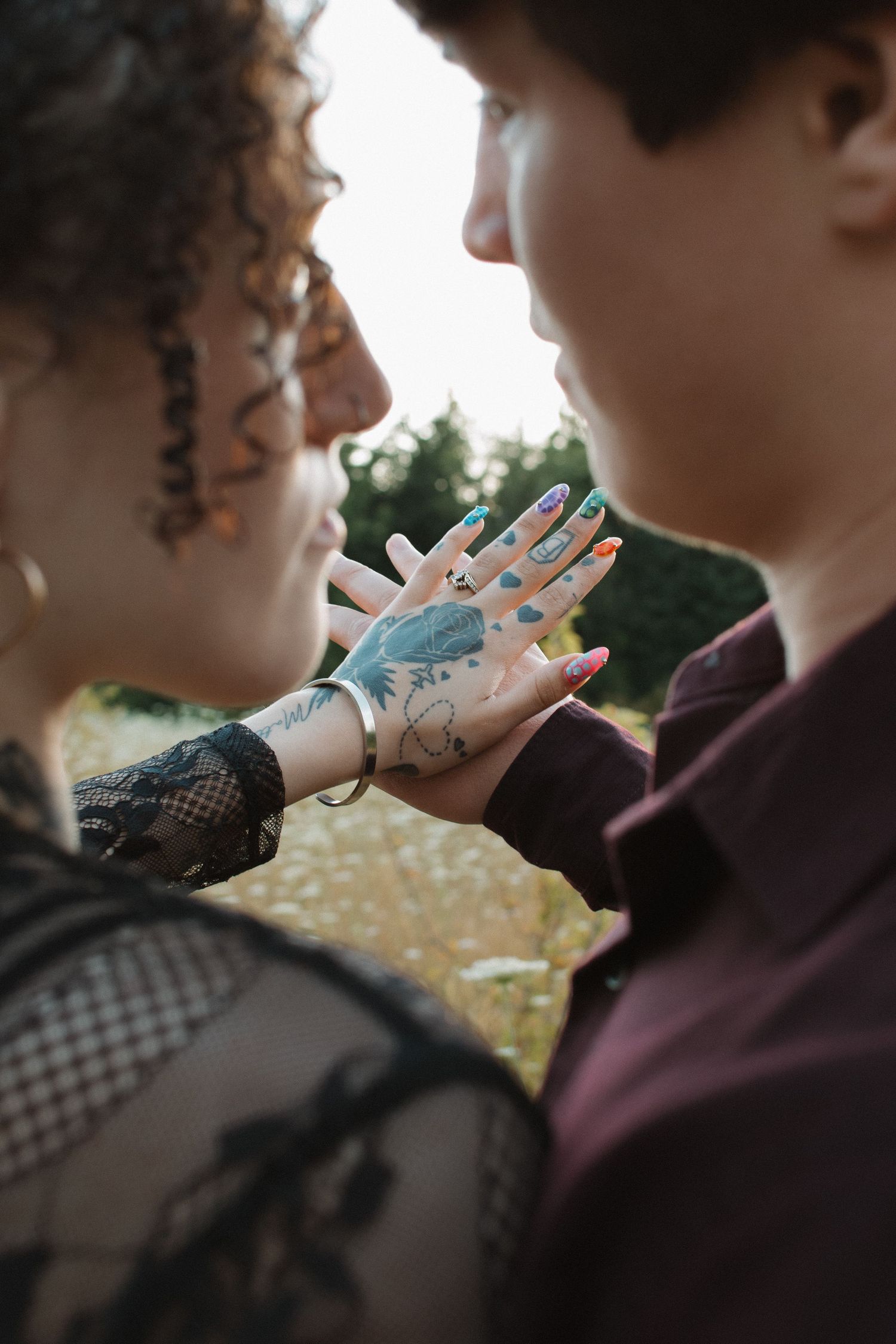 Couple holding tattooed hands together with colorful nail polish in outdoor natural setting.