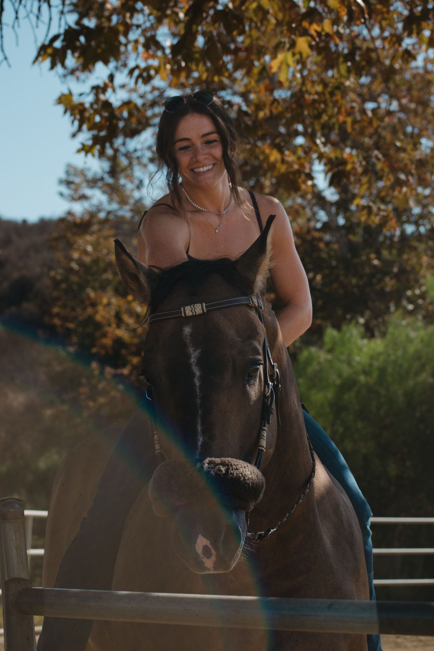 Smiling young woman riding a dark brown horse outdoors with autumn trees in the background.