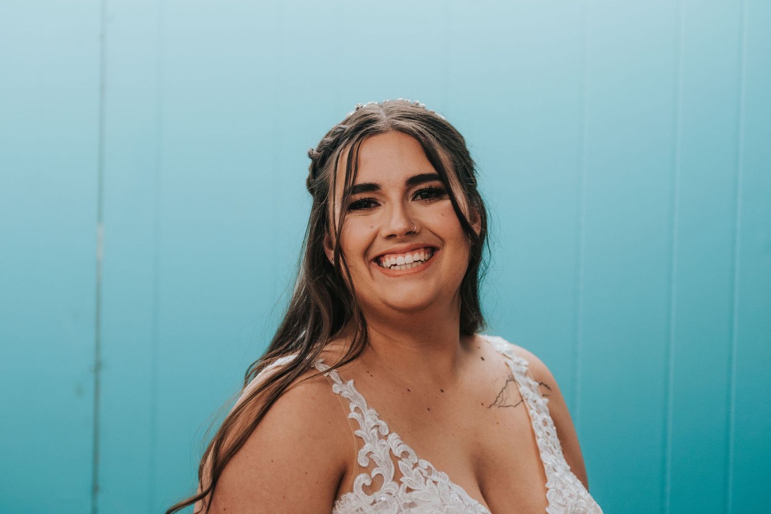 Smiling bride in lace wedding dress with floral crown against bright turquoise blue wall background.