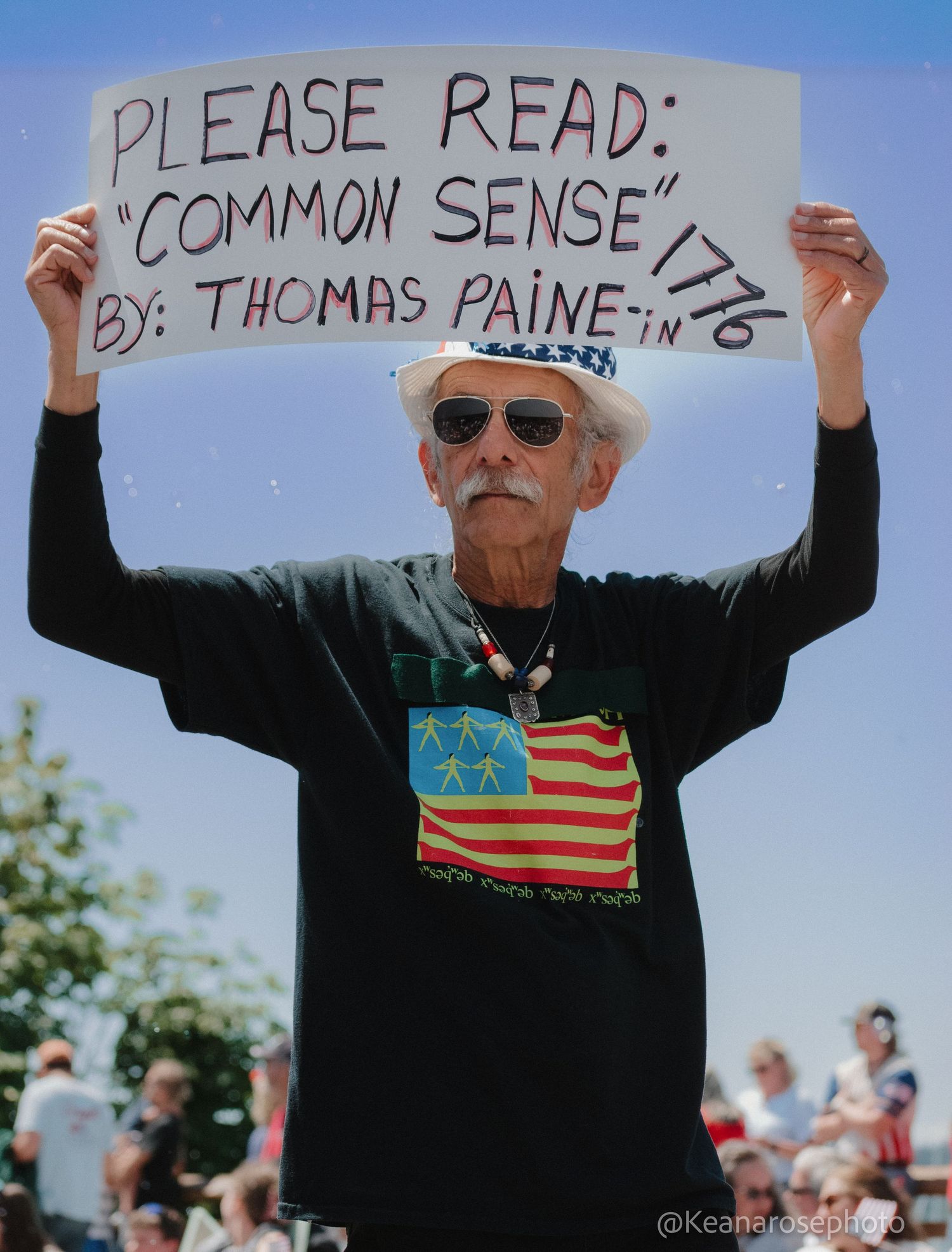 An older man holds a sign reading Please Read Common Sense 1776 by Thomas Paine at an outdoor rally.