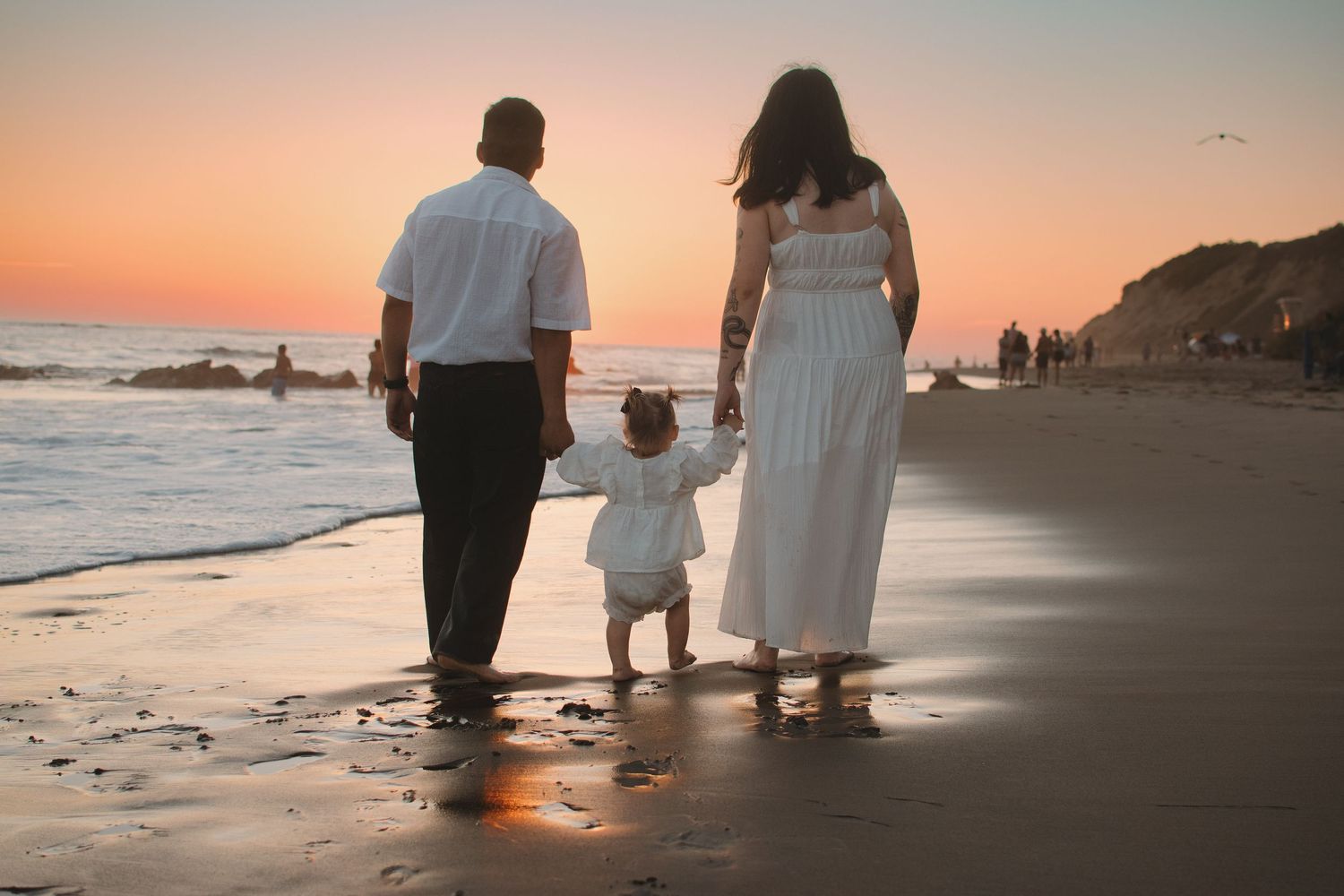 Family of three walking on beach at sunset, parents holding toddler's hands in matching white outfits.