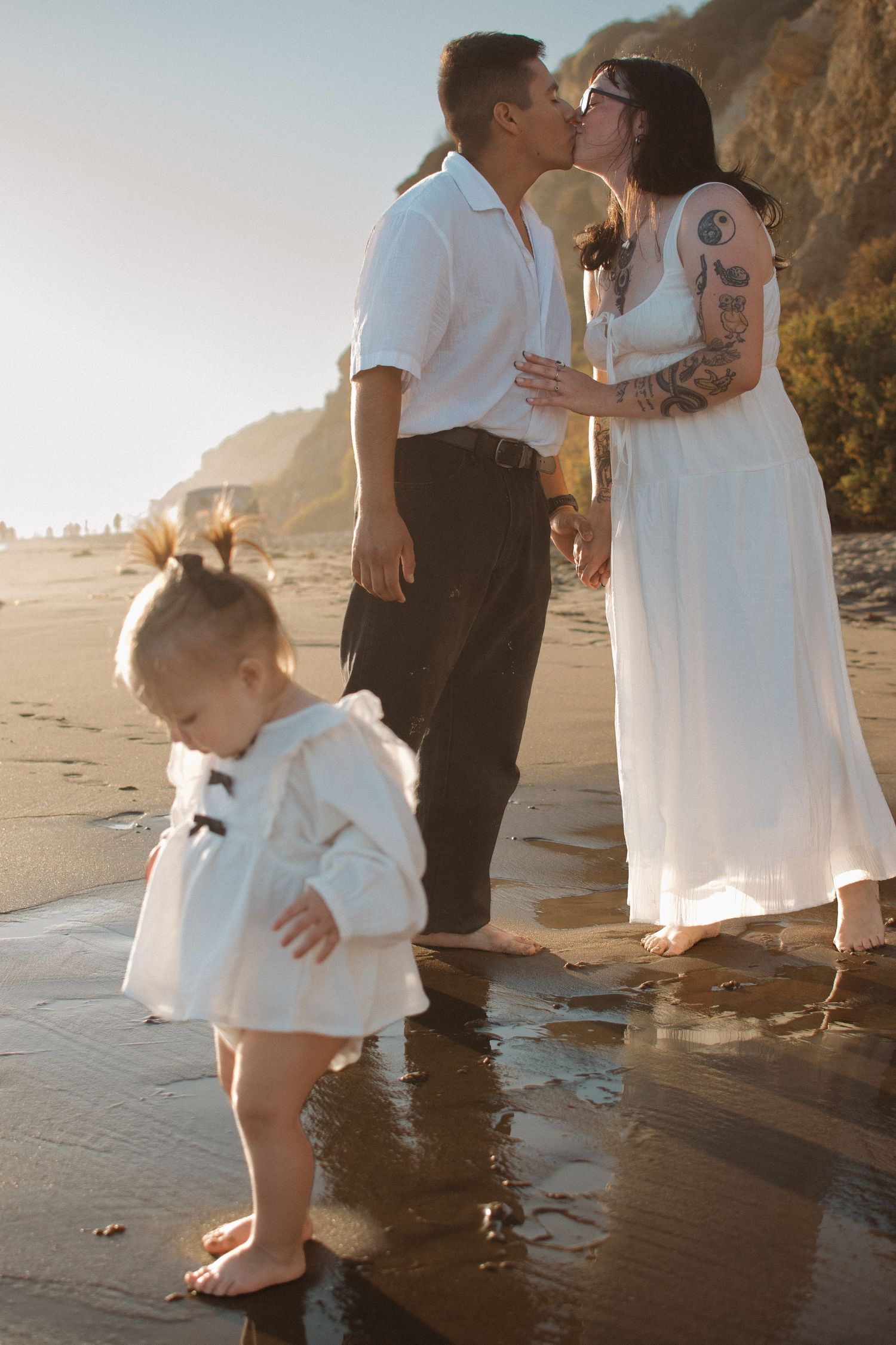 Couple kisses on a sunny beach while their young toddler daughter stands in the foreground.