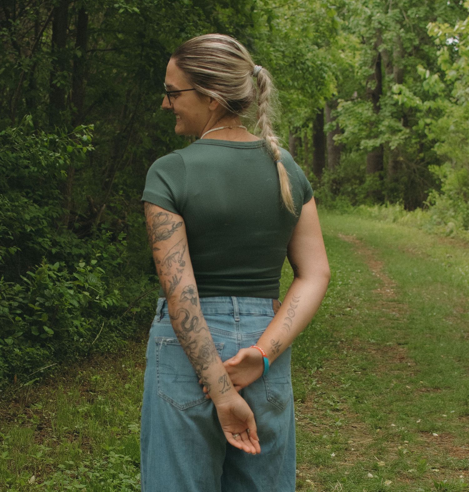 Woman with tattoos in green top and jeans stands on forest path, viewed from behind.