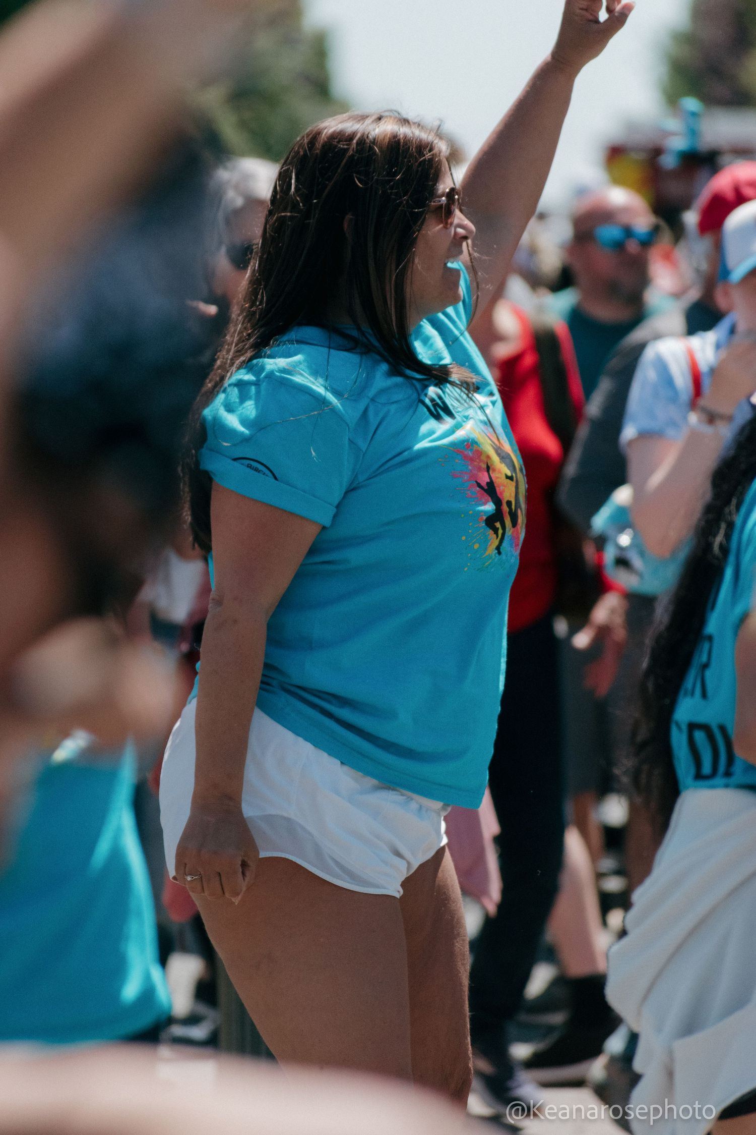 A woman in a blue t-shirt raises her fist in the air among a crowd at an outdoor public demonstration.