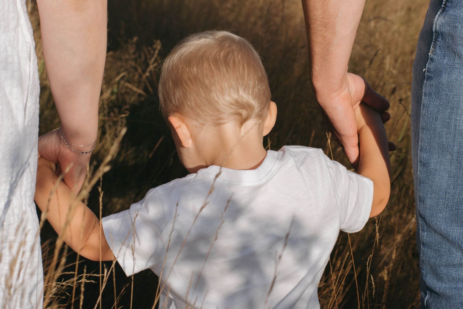 Young toddler in white shirt walking while holding both parents' hands in a golden wheat field.