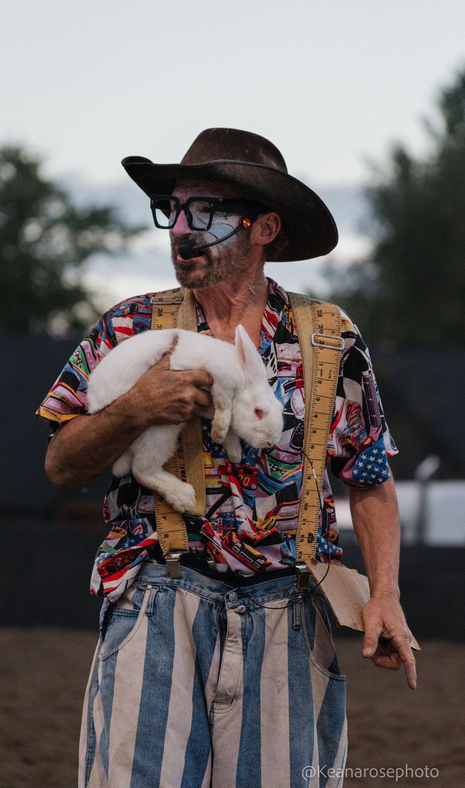 Rodeo clown in colorful outfit and face paint holds a white rabbit at a rodeo event.