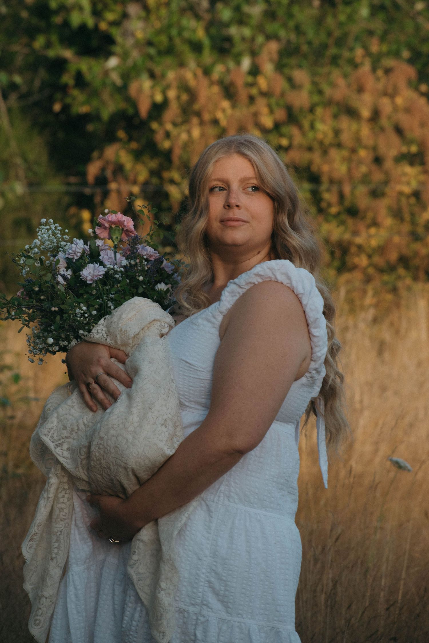 Blonde woman in white dress holding a bouquet of wildflowers in a golden field at sunset.