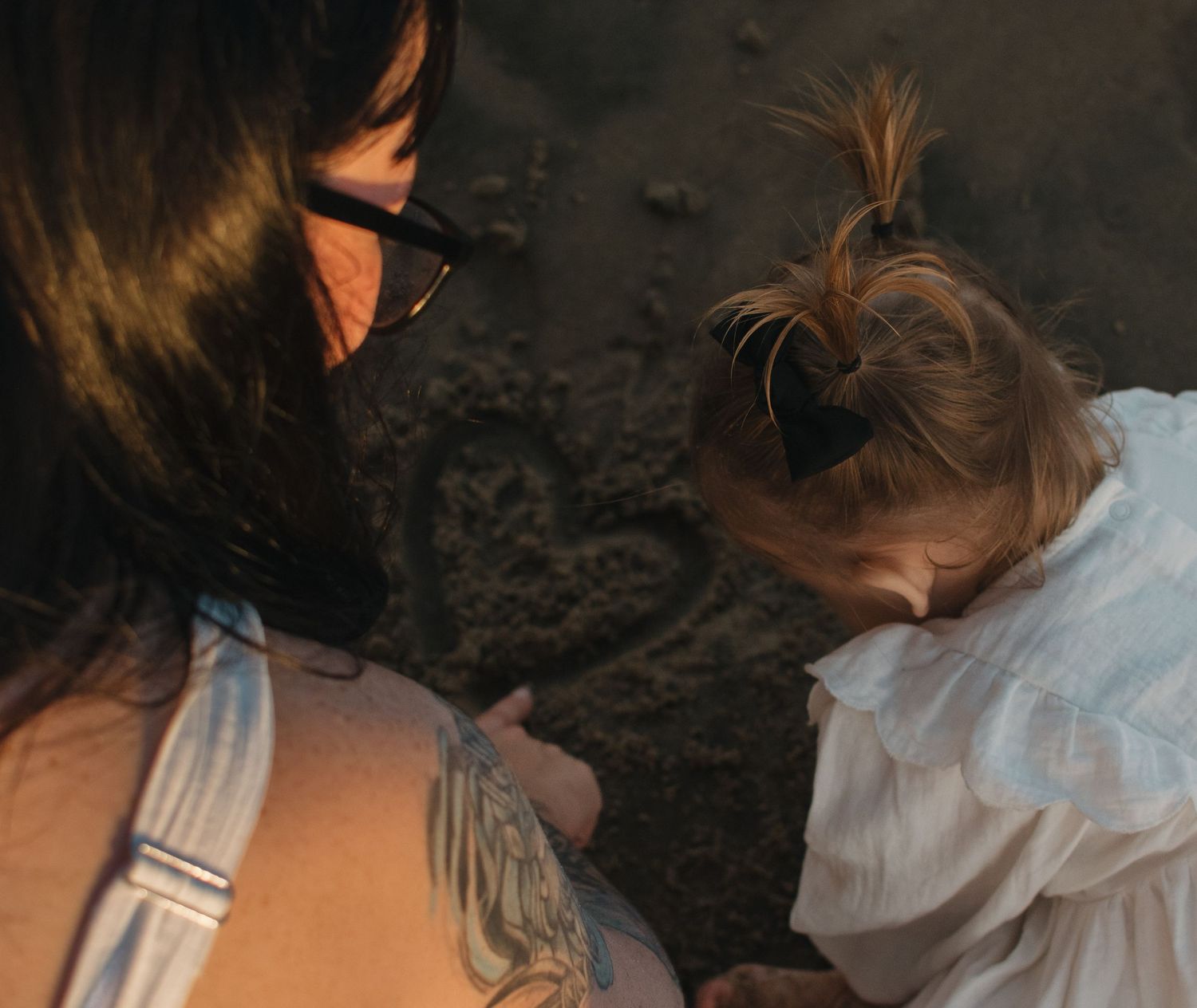 A tattooed woman with sunglasses and a young girl with a ponytail playing together in the sand.