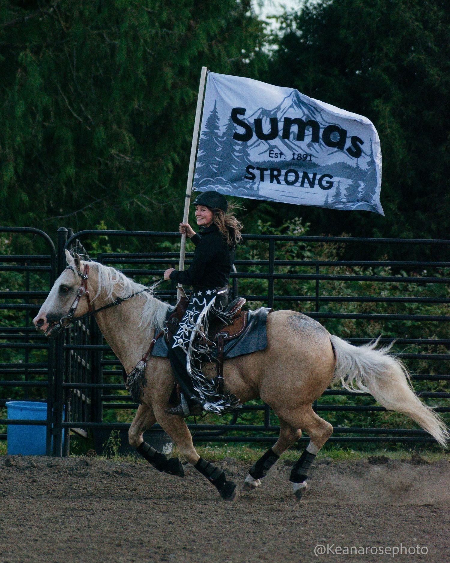 Female rider on palomino horse carries Sumas Strong flag while galloping in rodeo arena at dusk.