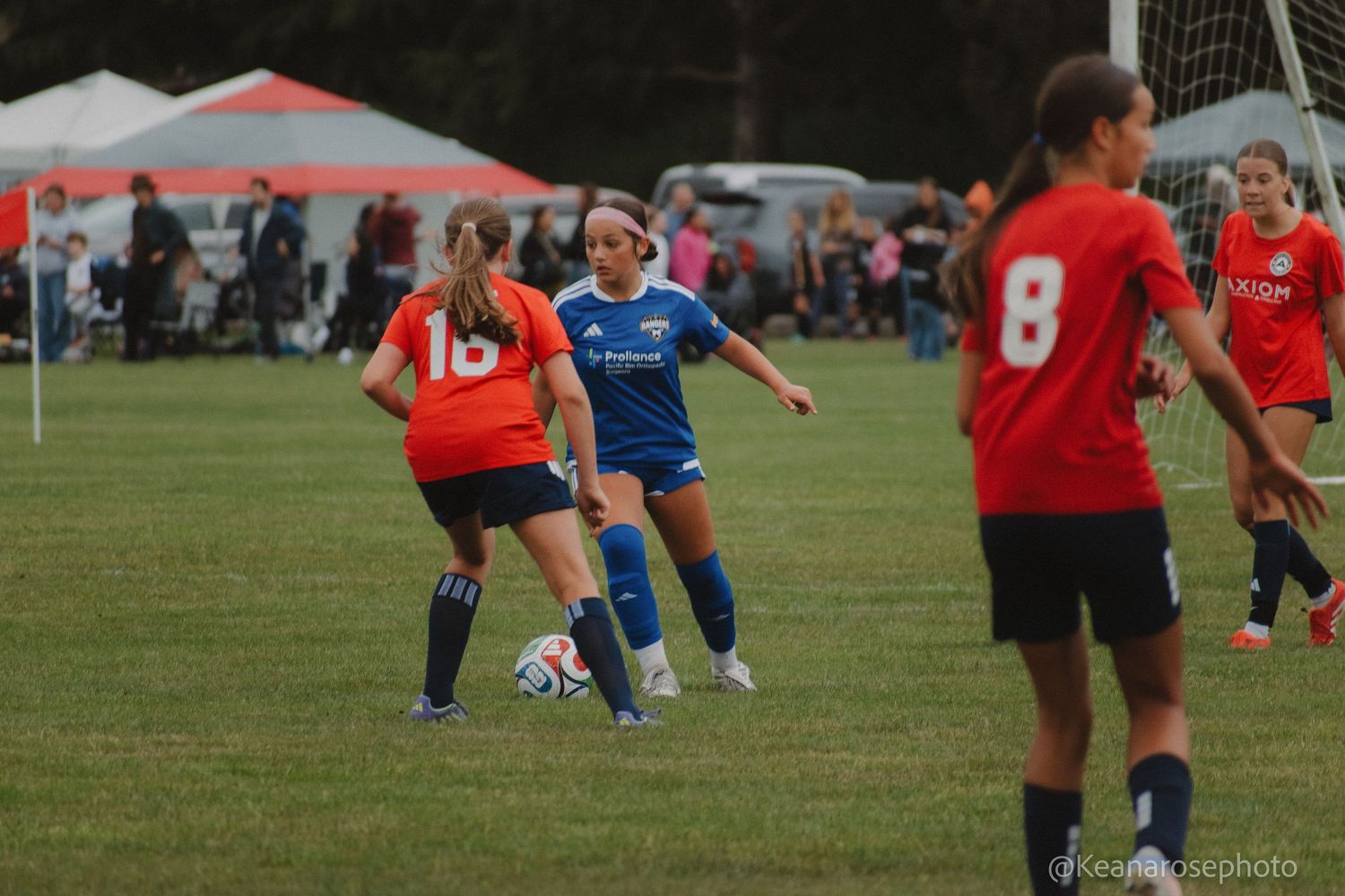 Female youth soccer players in red and blue uniforms compete for the ball during an outdoor match.