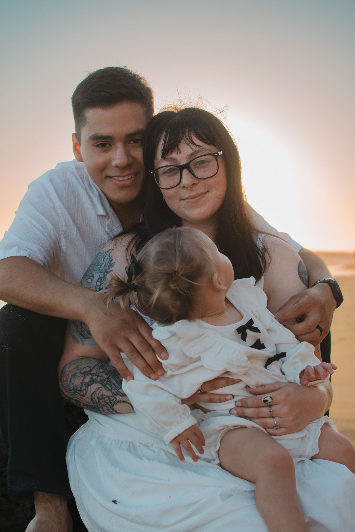 A young family with tattoos and glasses poses together at sunset on a beach, holding their baby girl in a white dress.