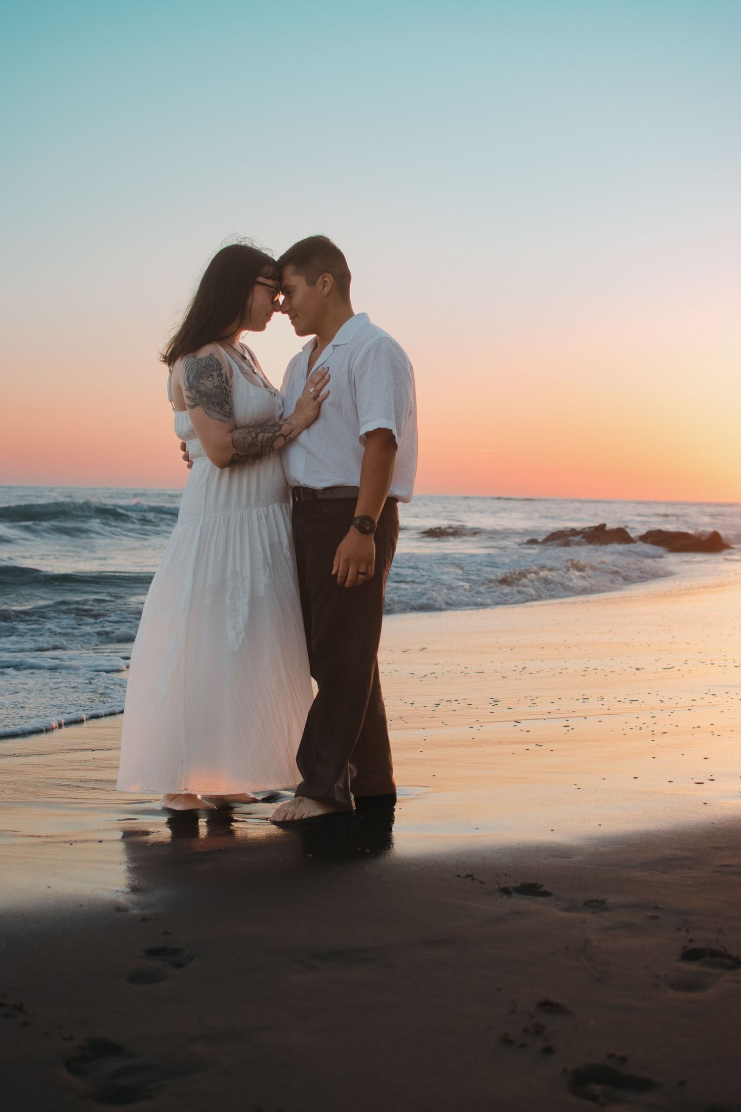 Romantic couple sharing an intimate moment on a sandy beach at sunset, waves and colorful sky in the background.