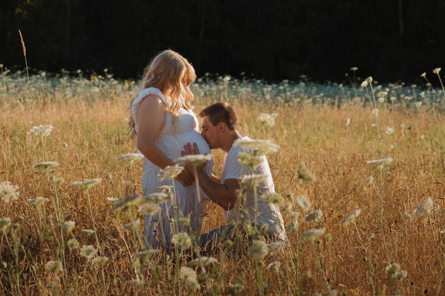 Man kneeling in golden wildflower field kissing pregnant woman's belly during outdoor maternity photo session.
