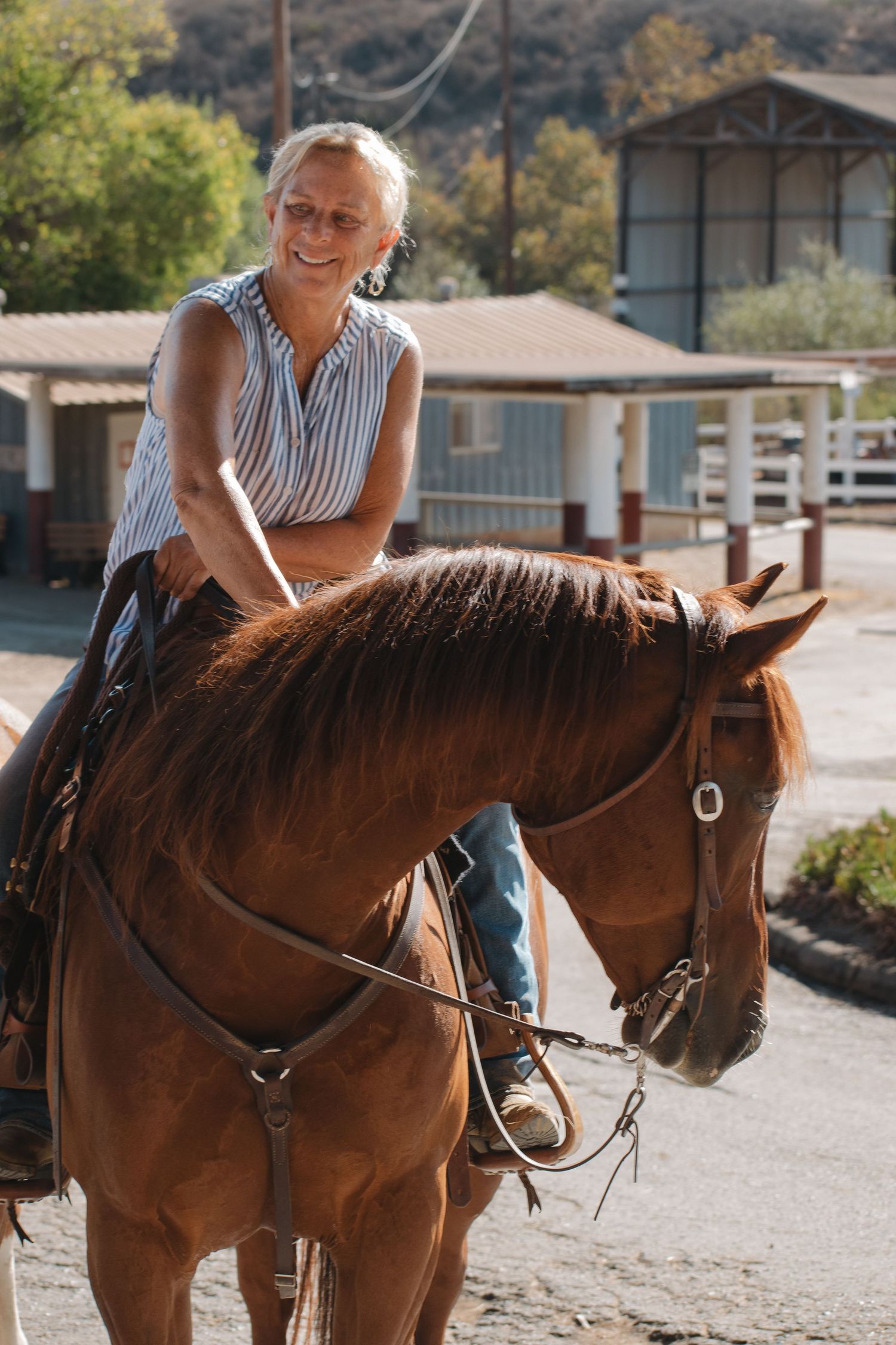 Smiling senior woman riding a chestnut horse at a sunny equestrian ranch facility.