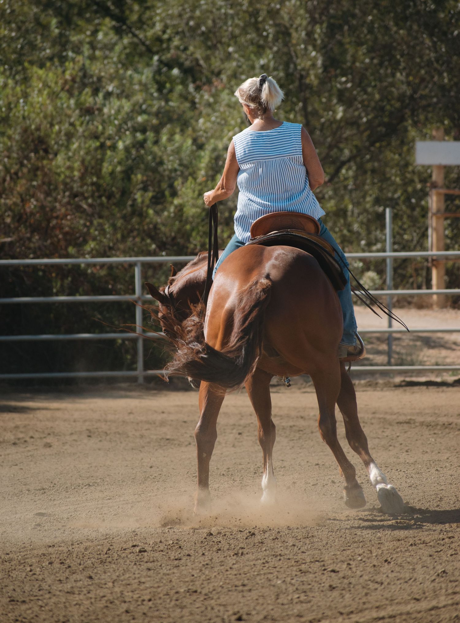 Blonde woman riding a chestnut horse in a dusty outdoor arena surrounded by trees.