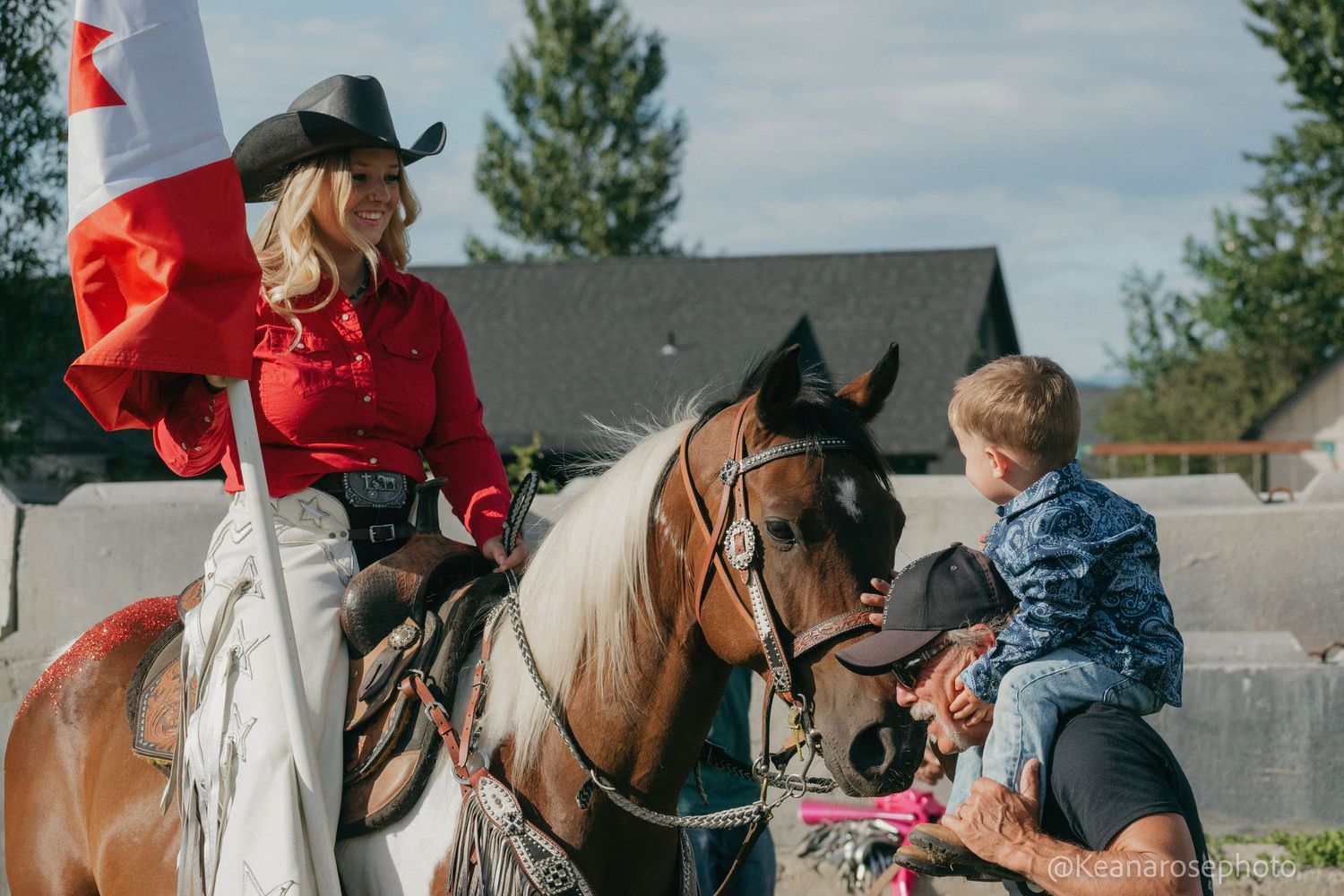 Blonde woman in red shirt on horseback holds Canadian flag while child touches horse at rodeo event.
