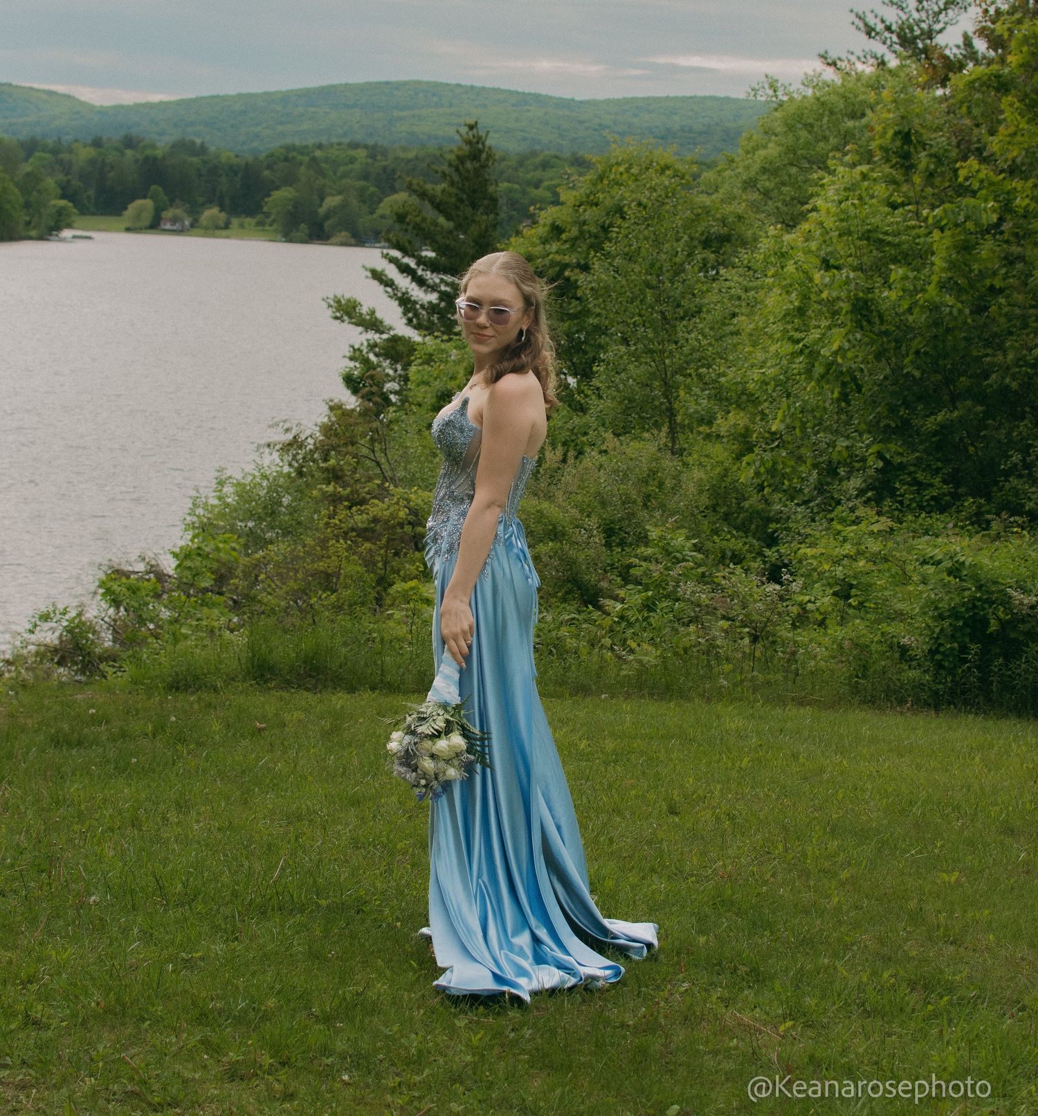 A young woman in a light blue prom gown holds a bouquet beside a serene lake surrounded by green trees.