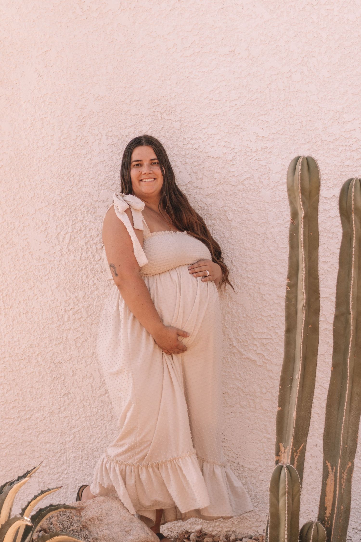 Smiling pregnant woman in cream ruffle dress holding belly, posing beside tall cactus against pink stucco wall.