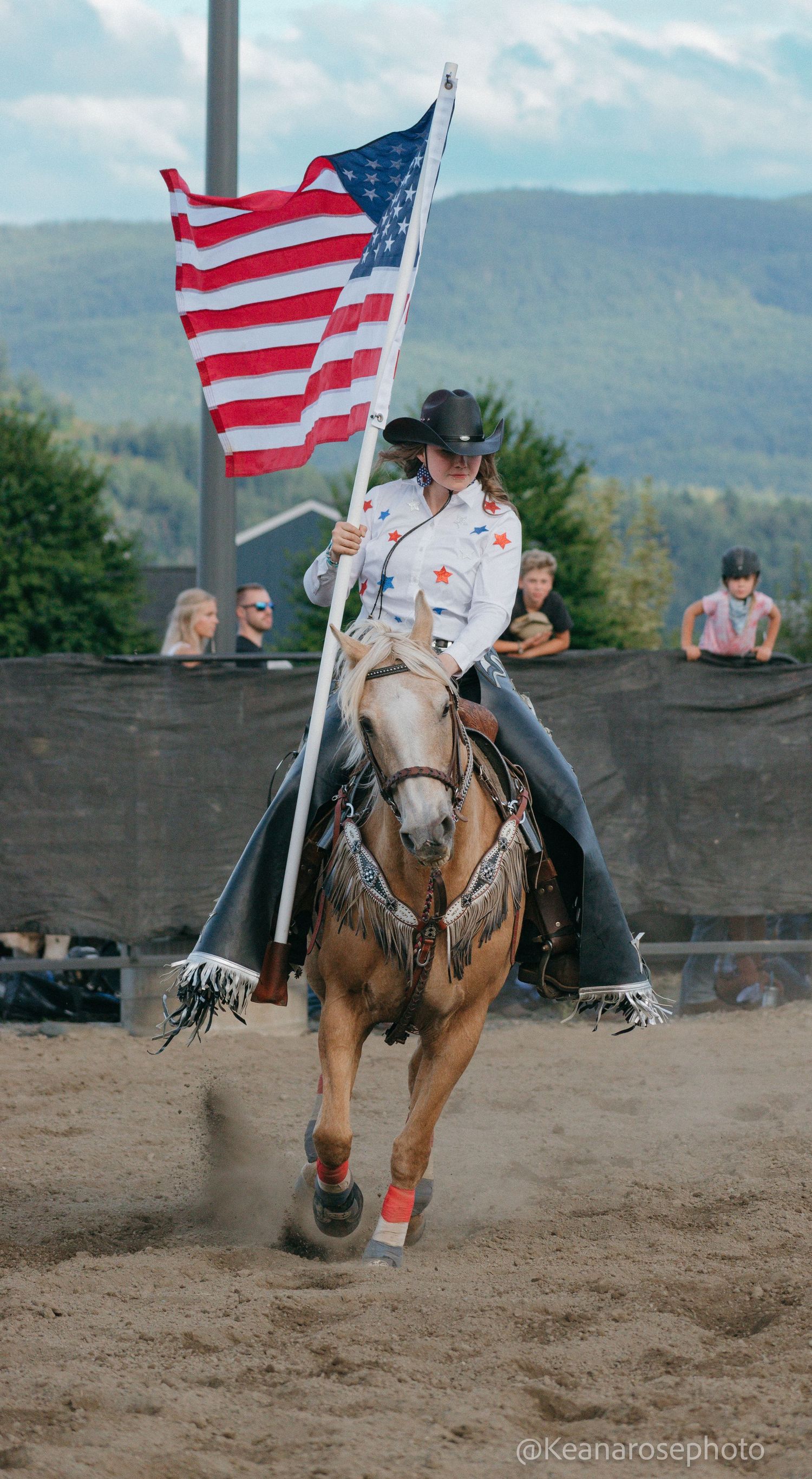 Female rider in white shirt carries American flag on palomino horse in mountain rodeo arena.