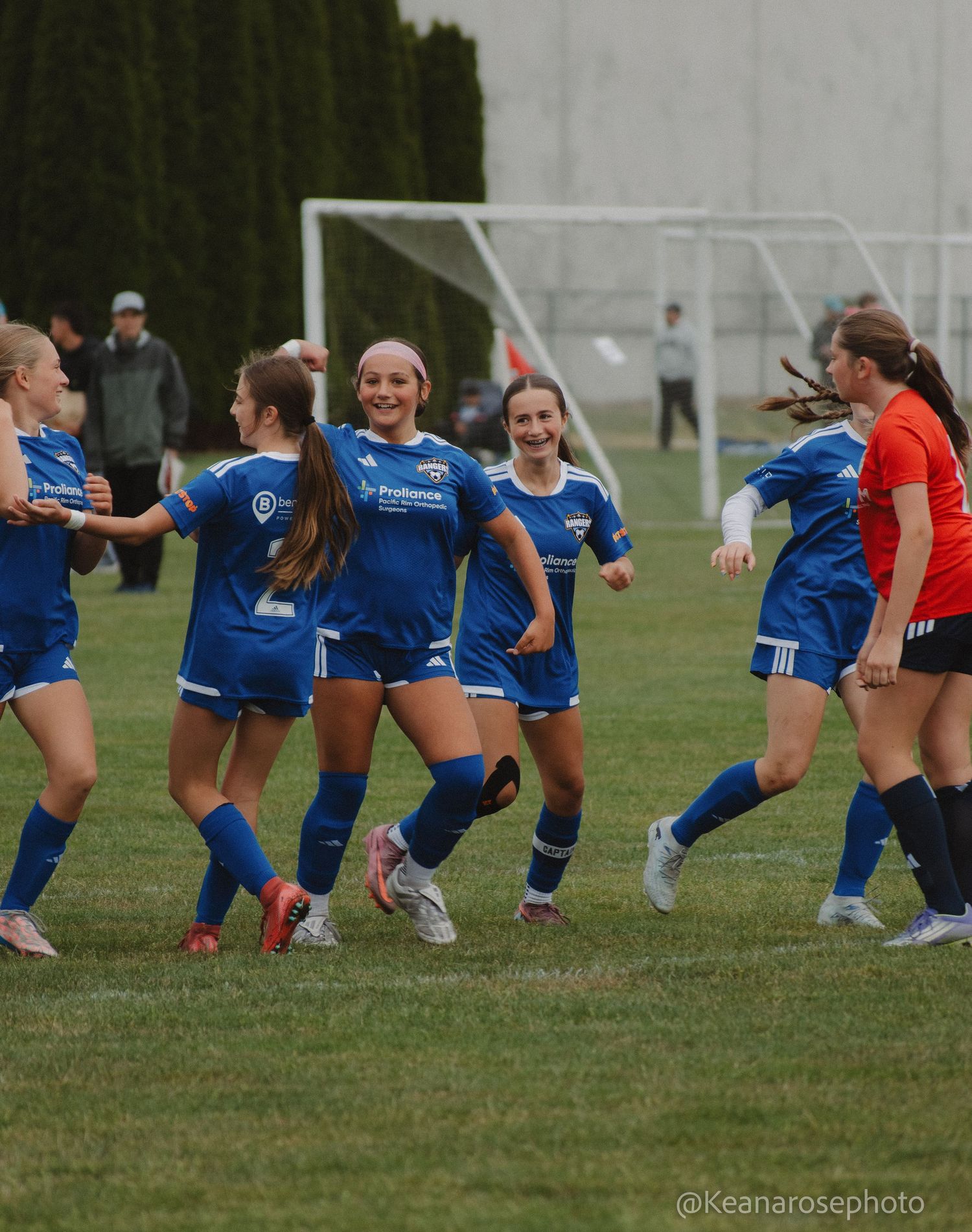 Girls youth soccer team in blue uniforms celebrate together smiling on the field after scoring a goal.