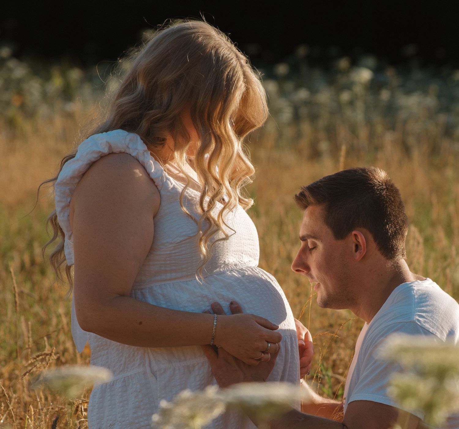 Pregnant woman in white dress with partner kneeling to kiss her baby bump in golden field.