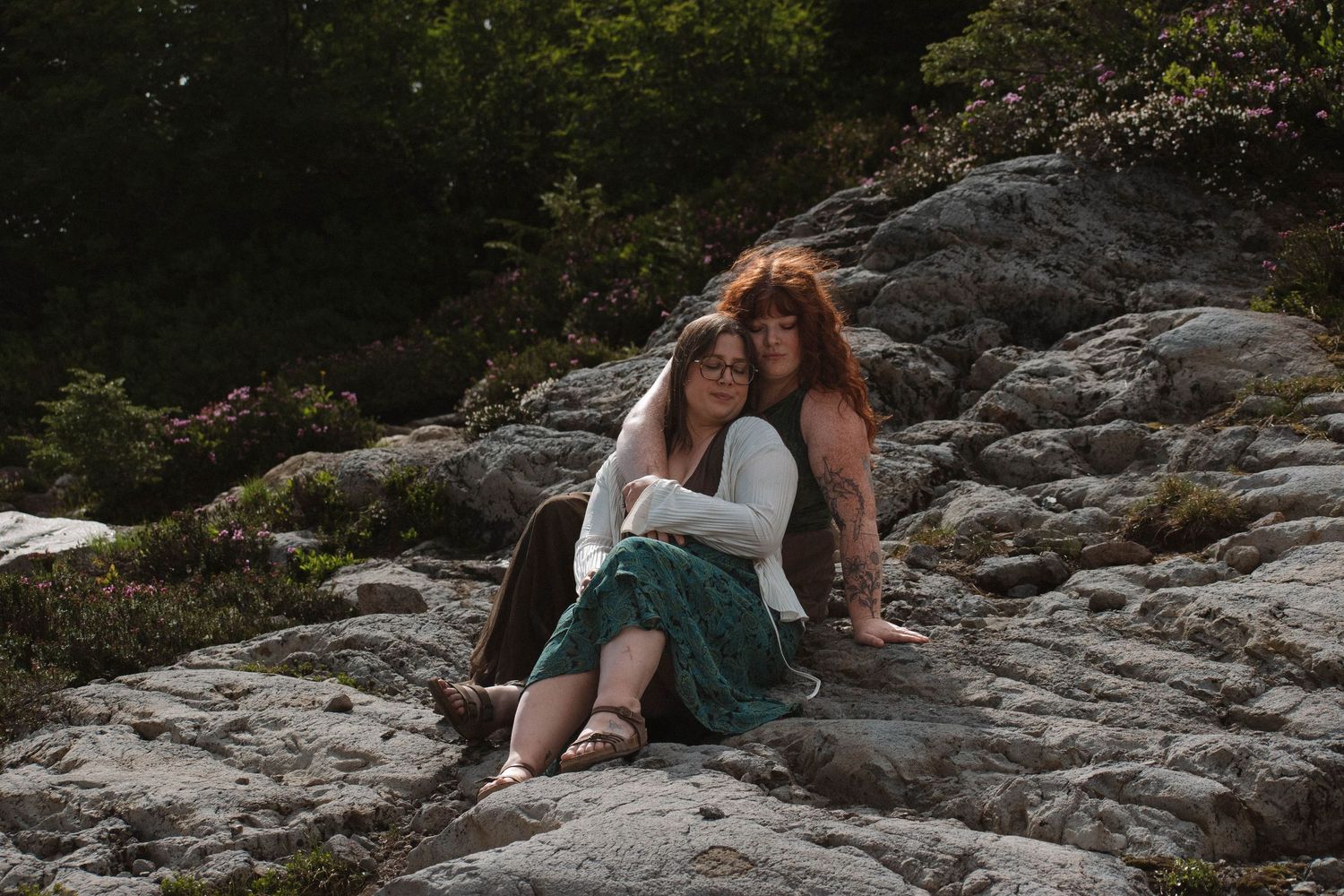 Two women sitting closely together on rocky terrain surrounded by lush green forest in outdoor portrait.