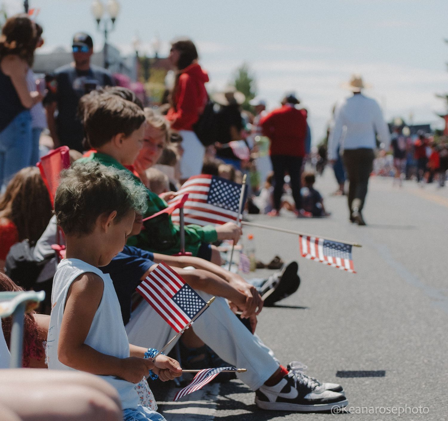Children sit along a sunny street waving small American flags while watching a Fourth of July parade.