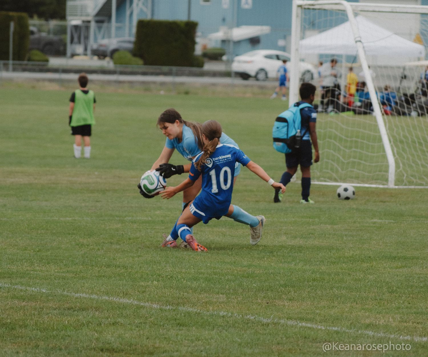 Youth soccer goalkeeper catches ball while player number 10 in blue slides in during a game.