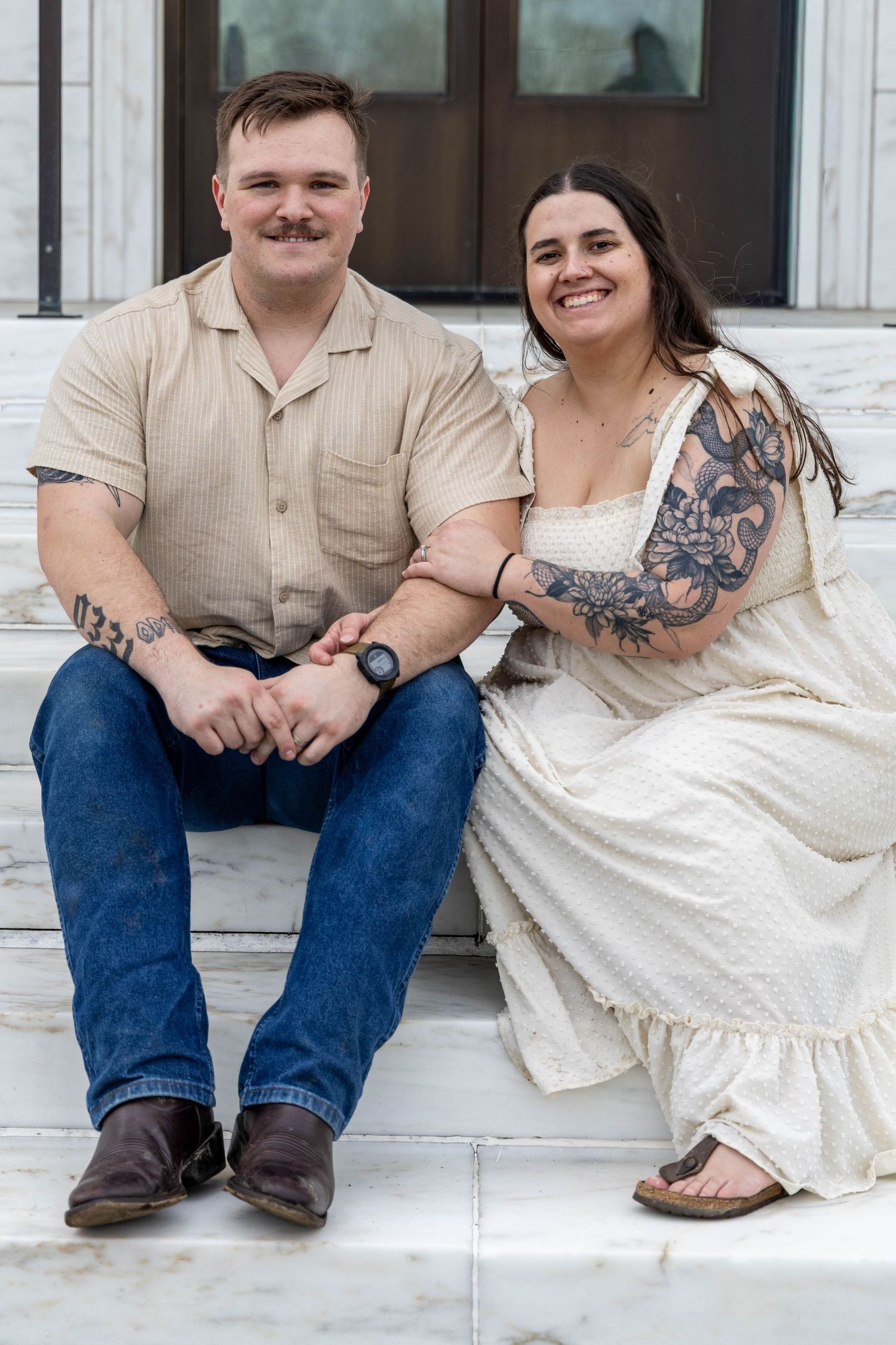 Tattooed couple sitting on marble steps smiling, man in beige shirt with jeans, woman in white dress.