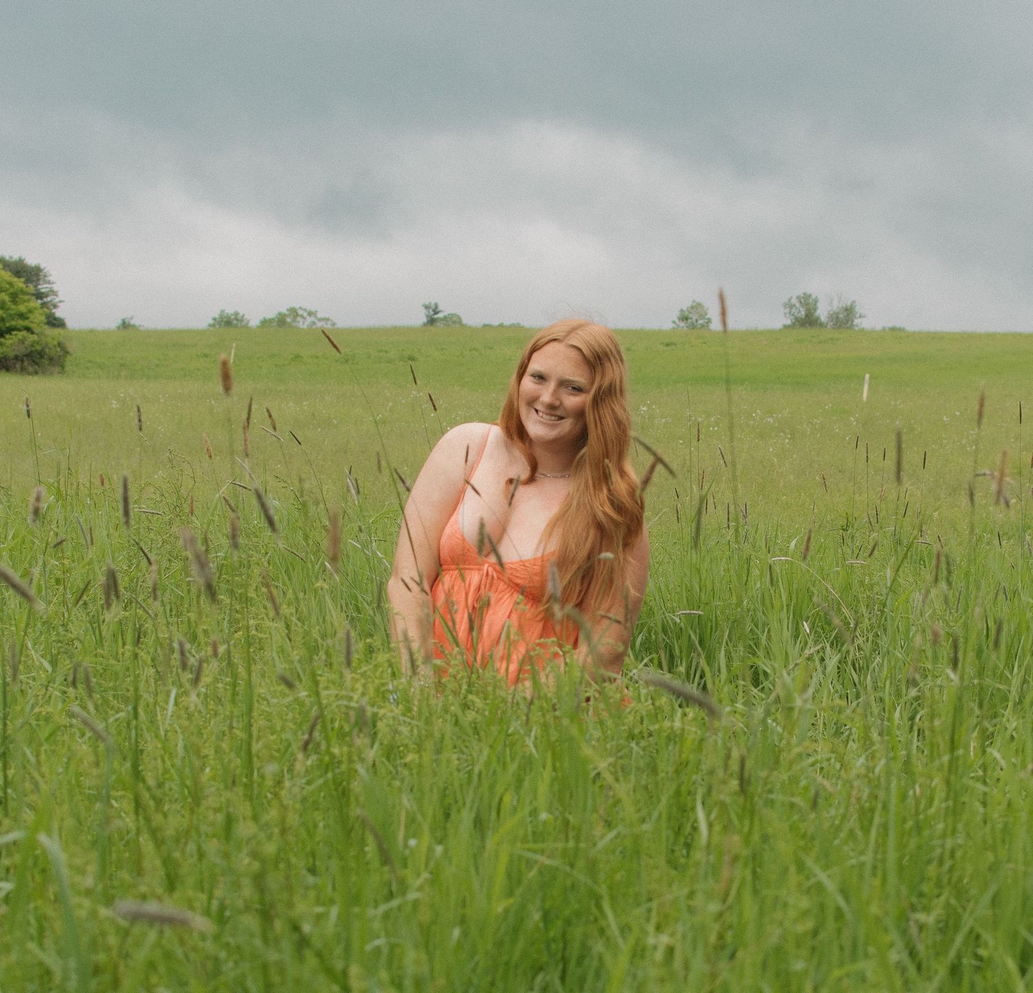 A smiling redhead woman in an orange dress sits in a lush green field under a cloudy sky.