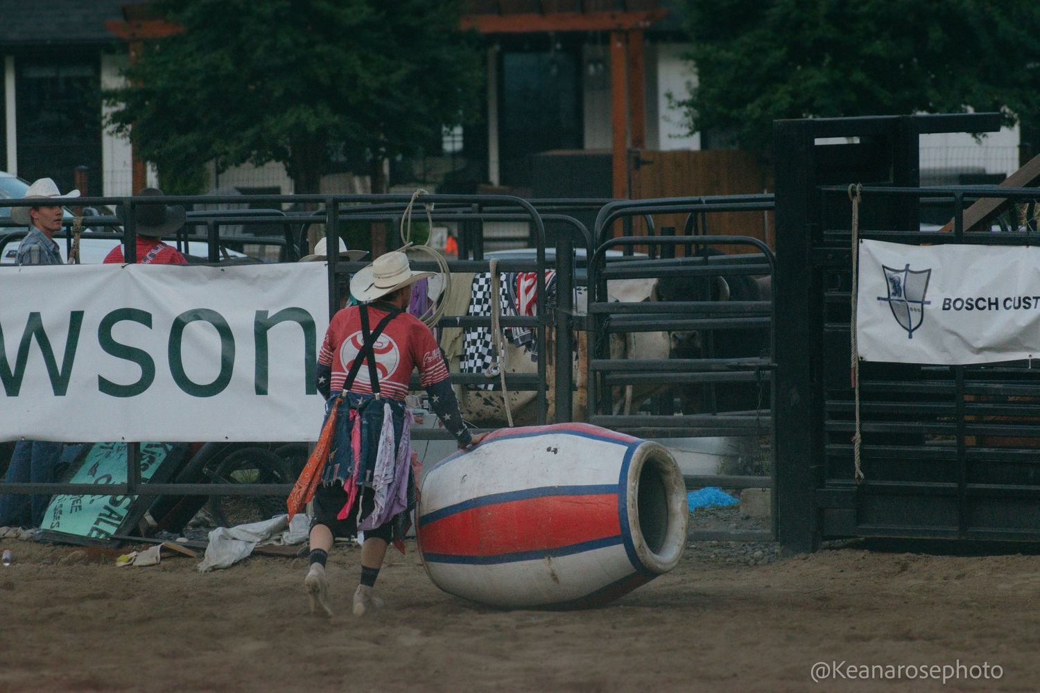 Rodeo clown in colorful costume stands next to a red white and blue barrel in the arena.