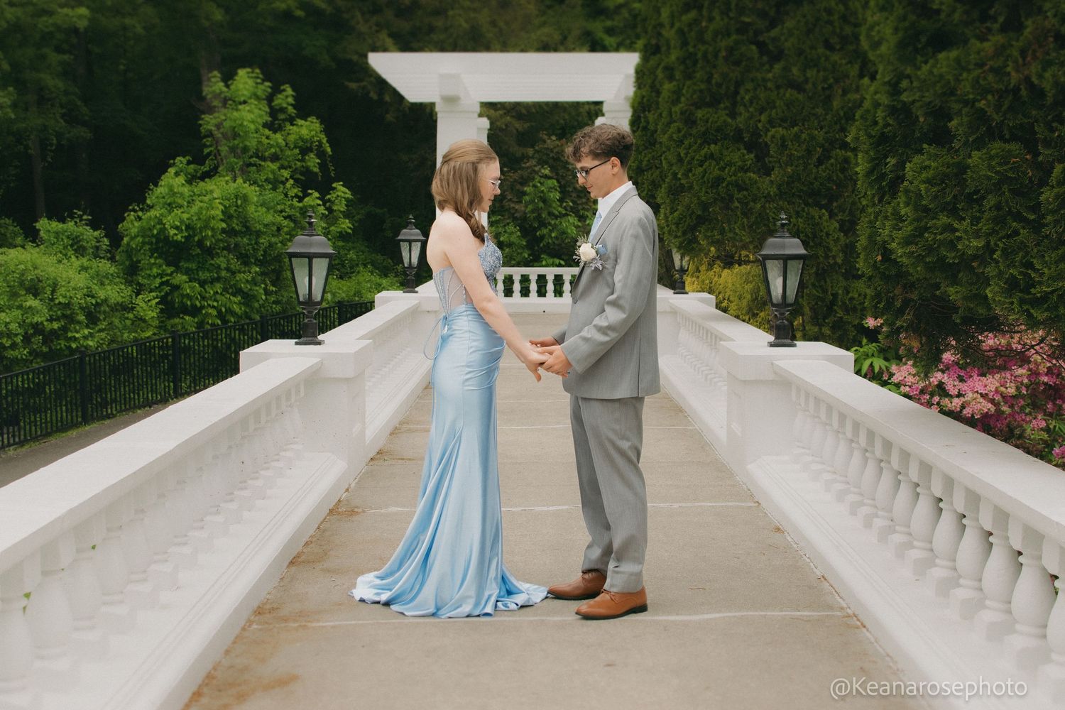 A couple in prom attire hold hands on a white balcony surrounded by lush green garden landscaping.