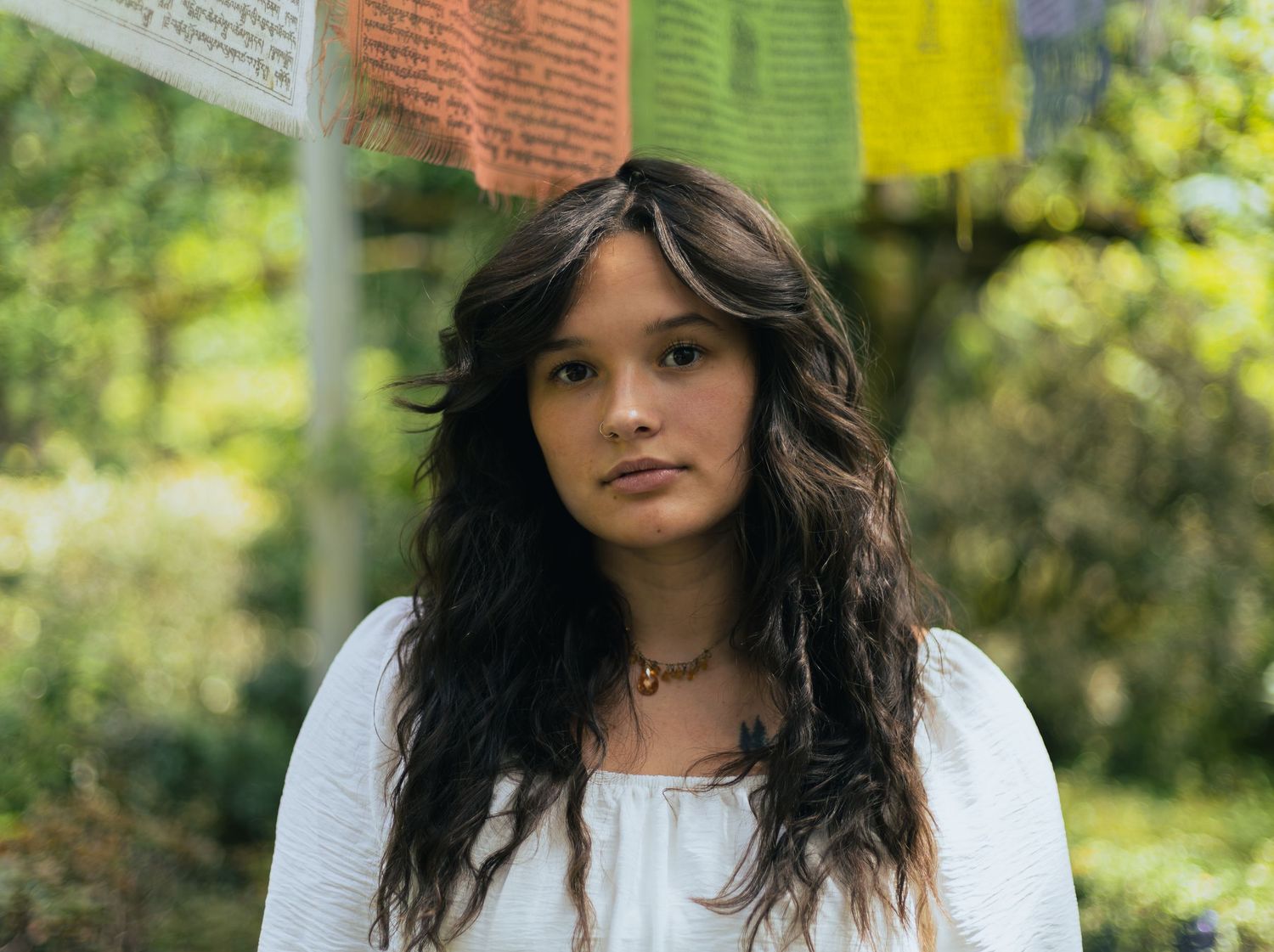 Young woman with long dark wavy hair wearing a white top standing outdoors near colorful prayer flags.