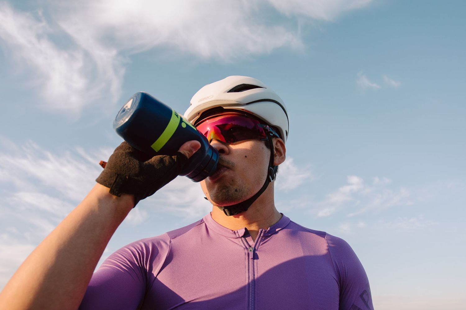 Male cyclist in purple jersey and white helmet drinks from water bottle against blue sky.