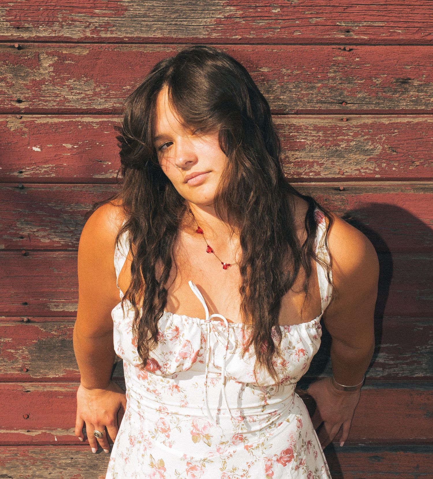 A young woman with long wavy brown hair wearing a white floral dress poses against a rustic red barn wall.
