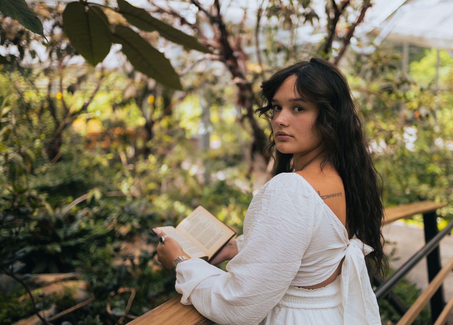 Young woman in white backless top holds open book while looking back in a greenhouse.