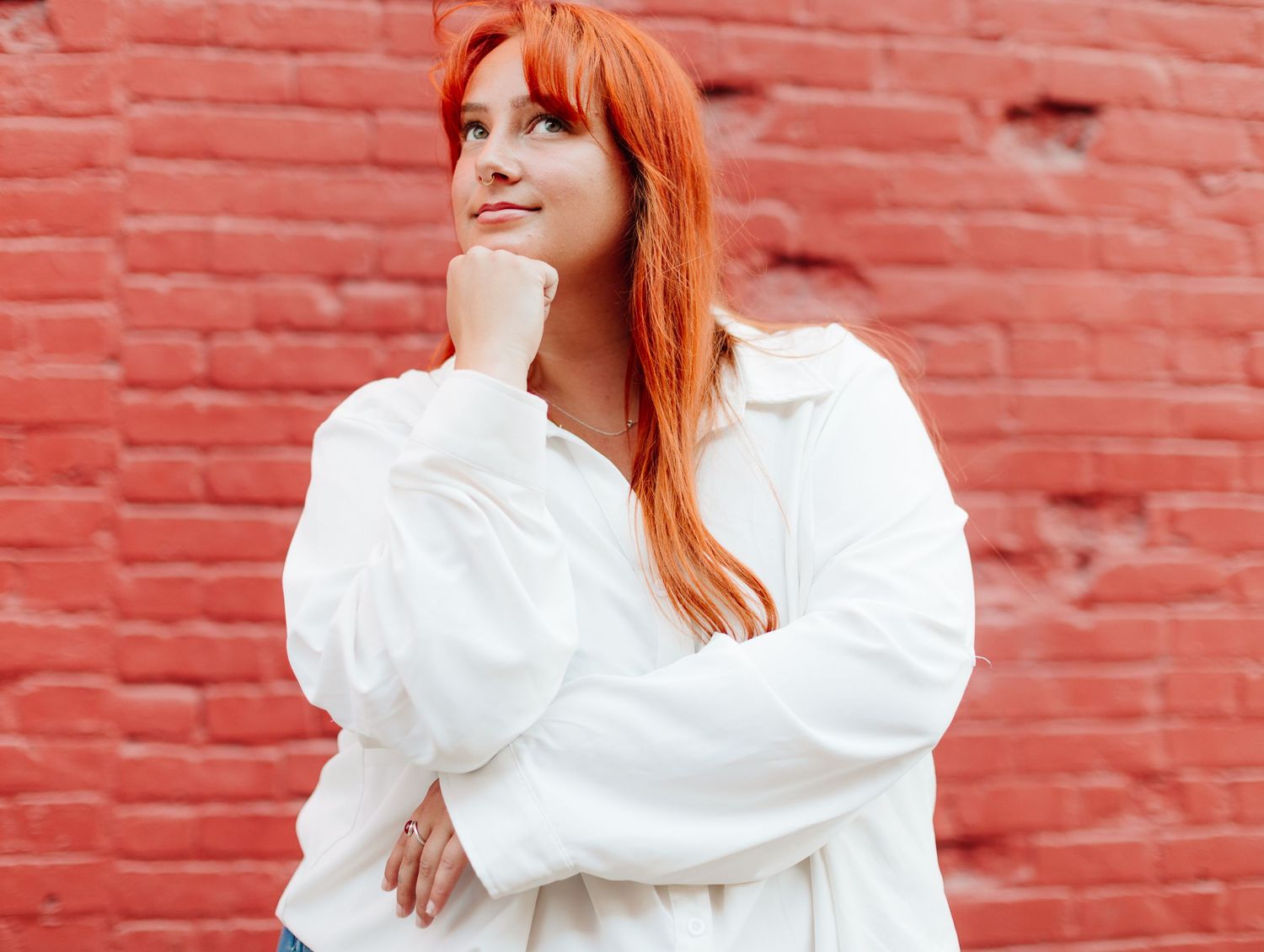Woman with bright red hair wearing white shirt posing thoughtfully against red brick wall.