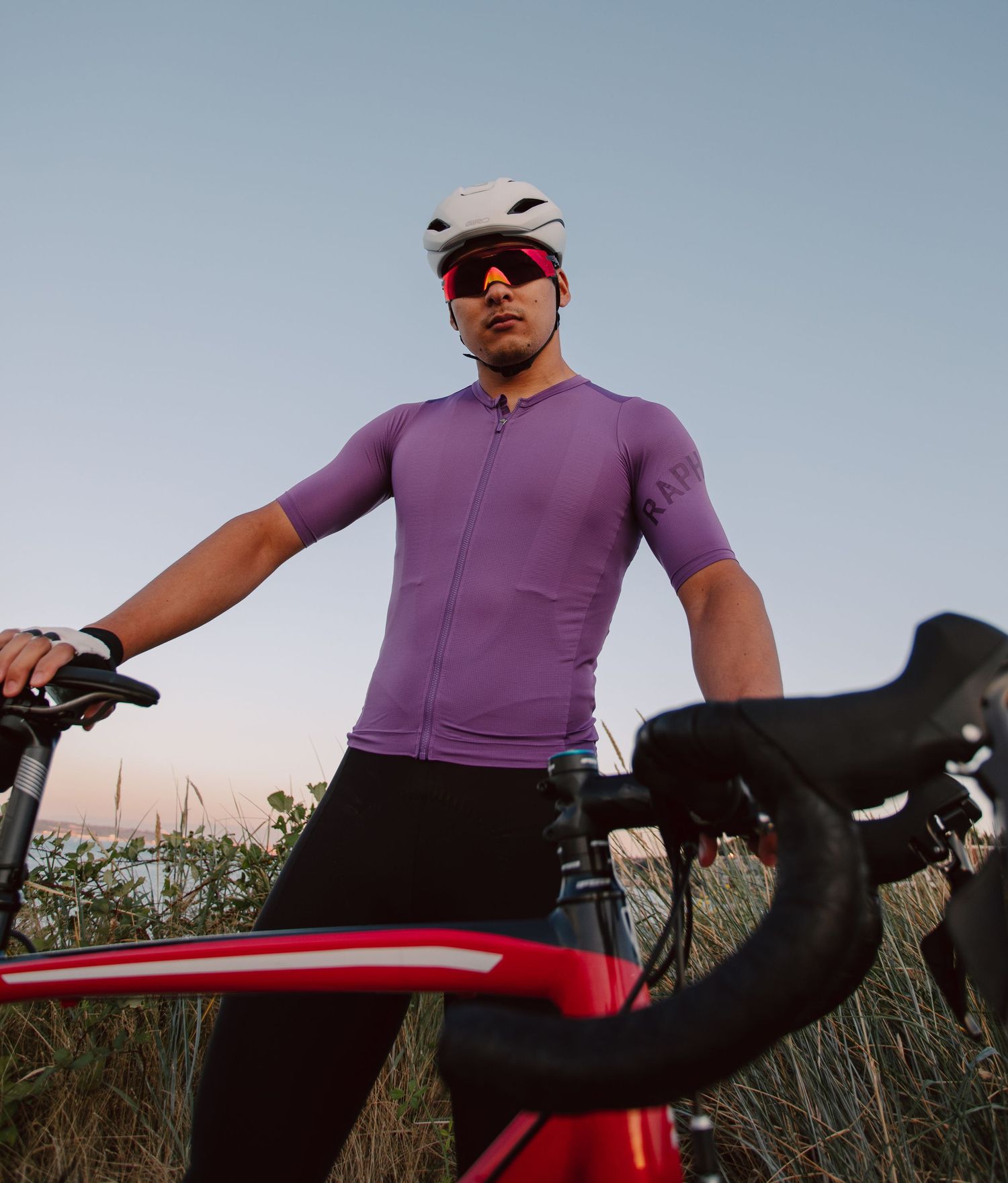 Male cyclist in purple jersey and white helmet stands with red Trek bike at dusk.